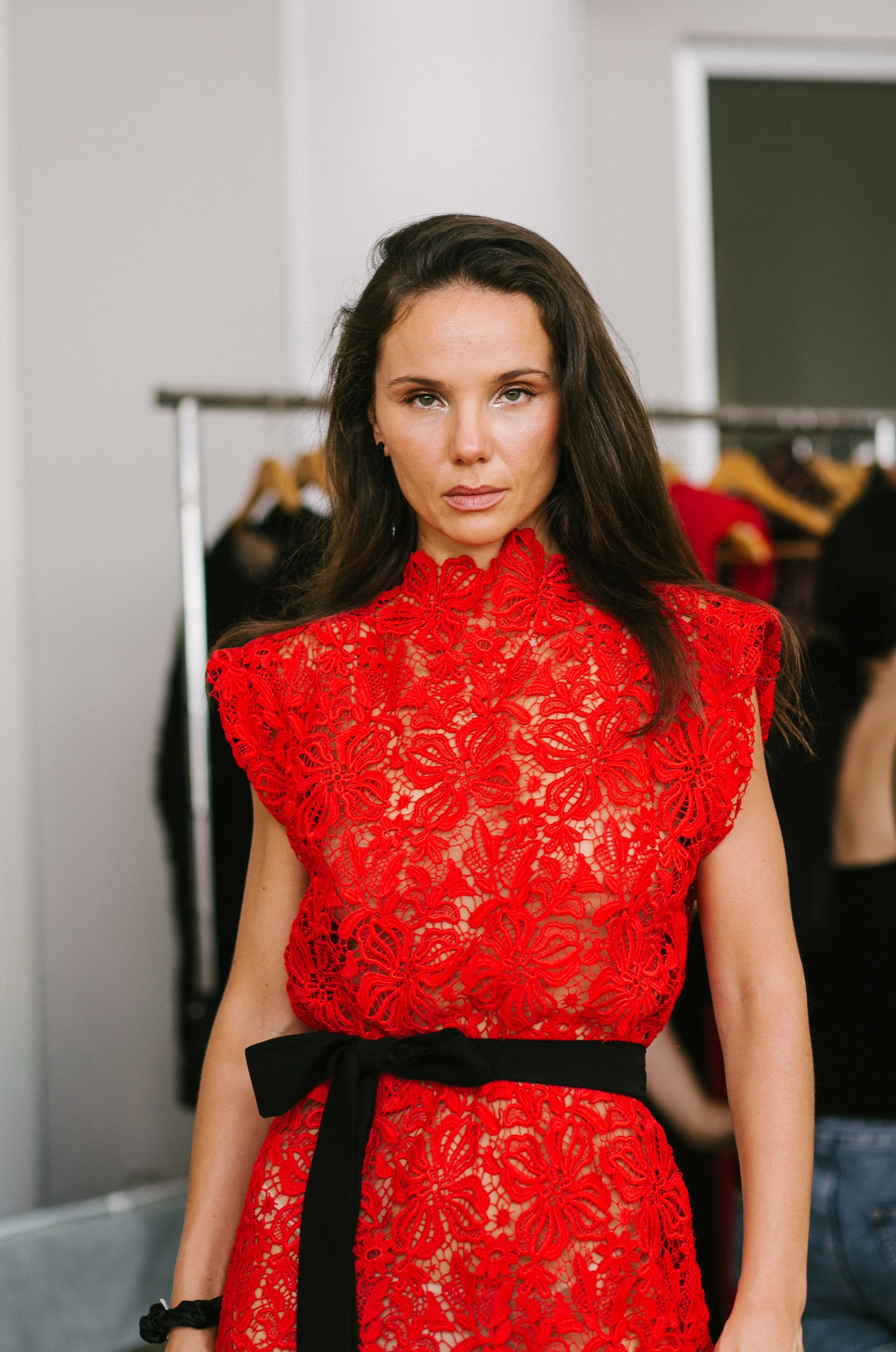 Woman with brown hair wearing a red lace dress with a black belt, standing in a room with clothing racks in the background.