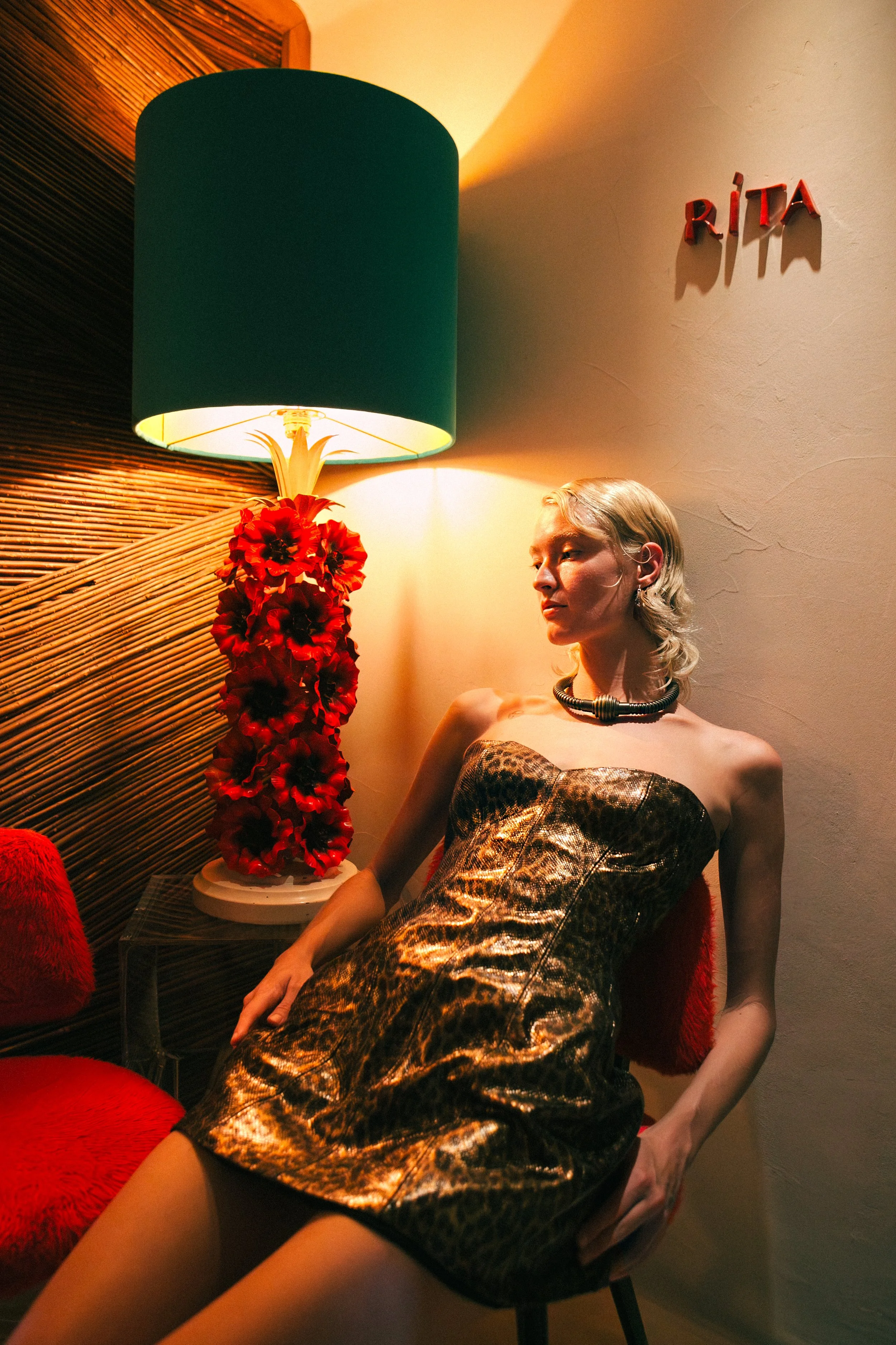A woman with blonde hair sitting in a red velvet chair next to a table with a tall lamp with a green shade and red flower base, in a room with warm lighting. The wall behind her has letters spelling 'RITA'.