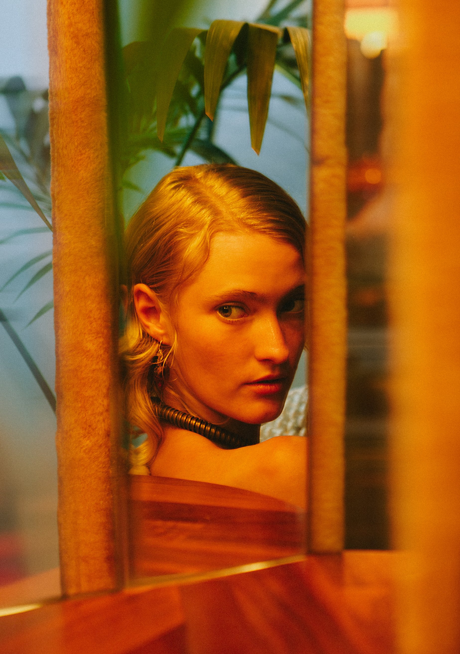 A woman with blonde hair looking over her shoulder through a mirror, surrounded by green plants and warm lighting.