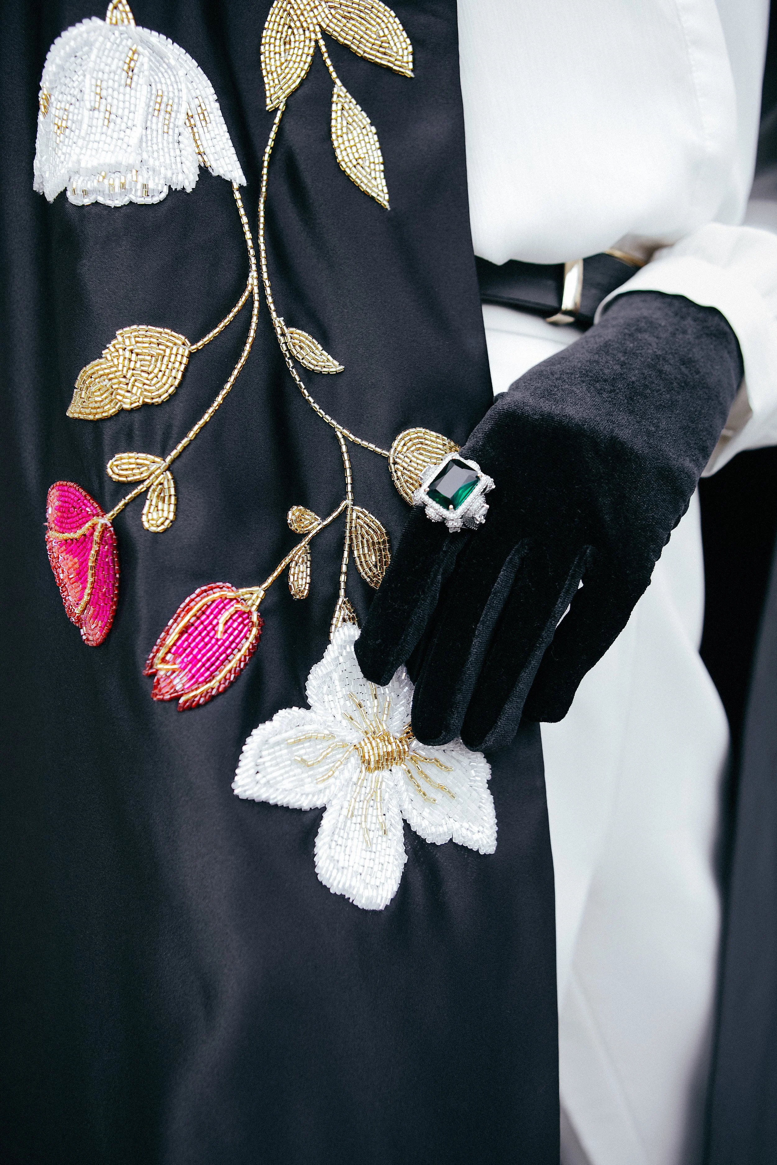 Close-up of a beaded floral brooch worn with black gloves, and a black and white garment, featuring pink and white embroidered flowers.