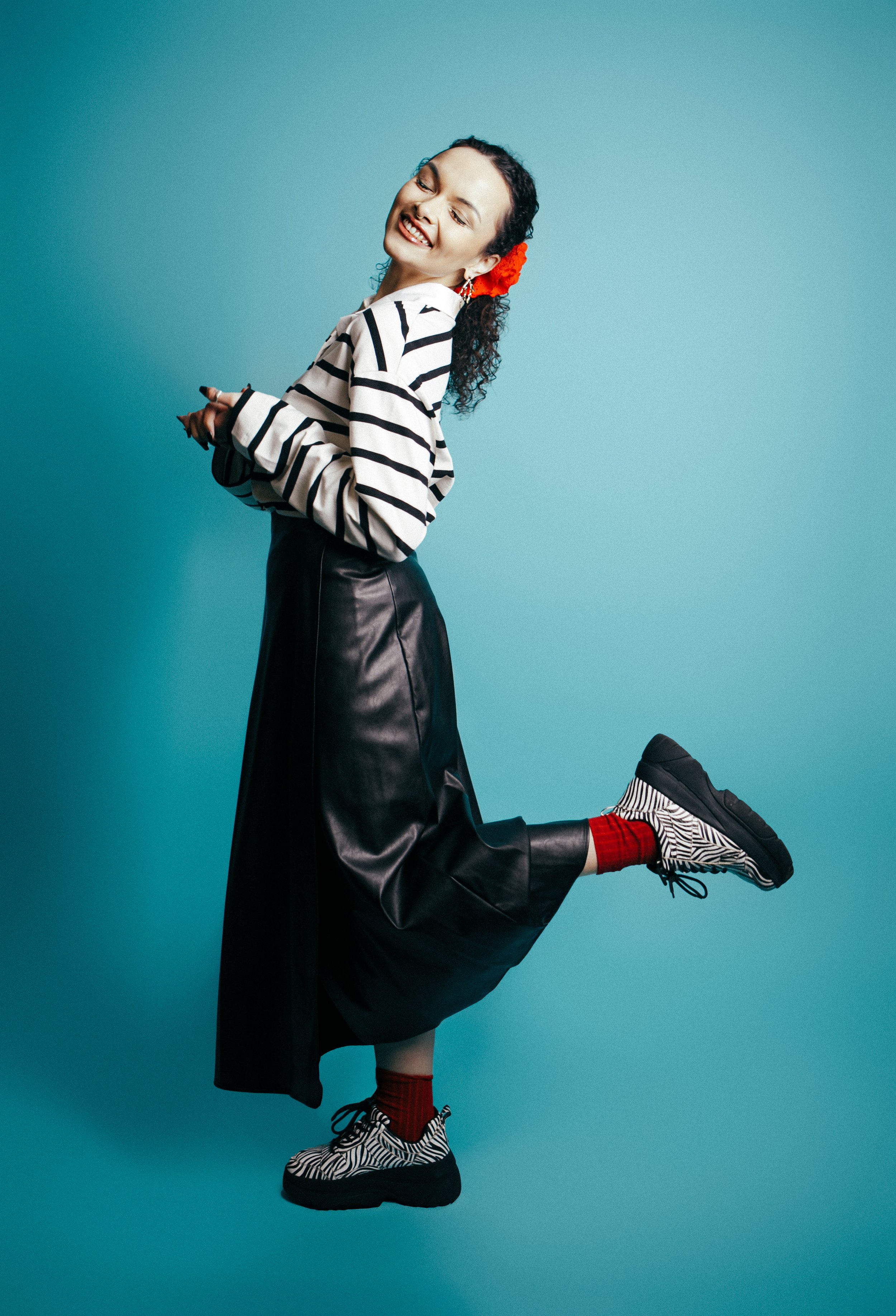 A woman with curly hair, wearing a striped shirt, long black leather skirt, zebra print sneakers, red socks, and a red hair accessory, smiling and posing against a solid teal background.