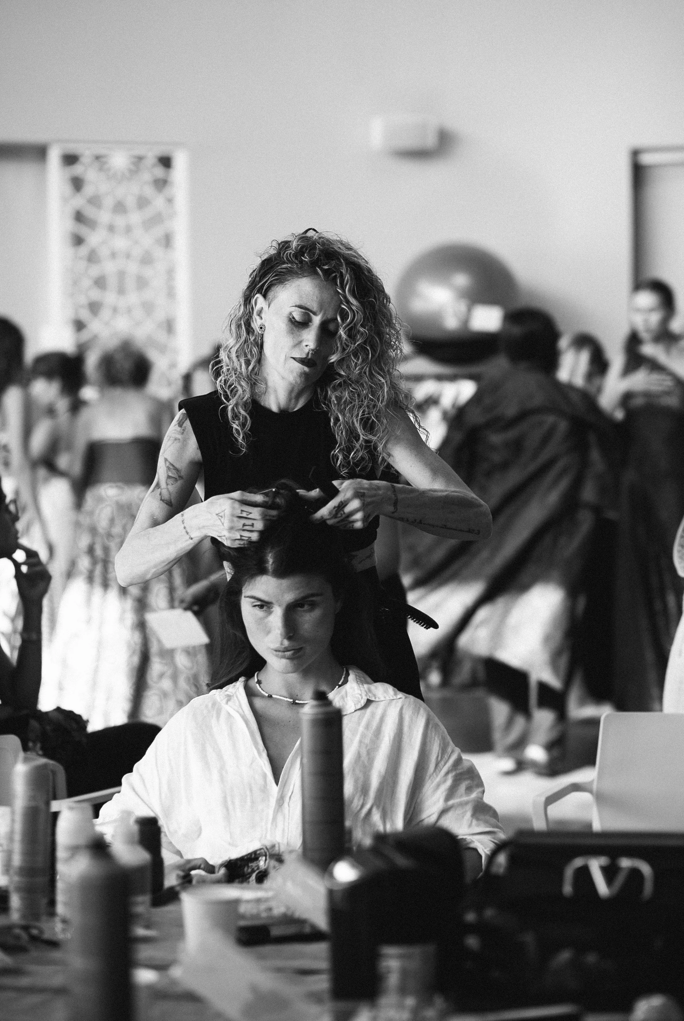 A woman with curly hair and tattoos is styling a woman's hair at a busy backstage area with people and fashion in the background.
