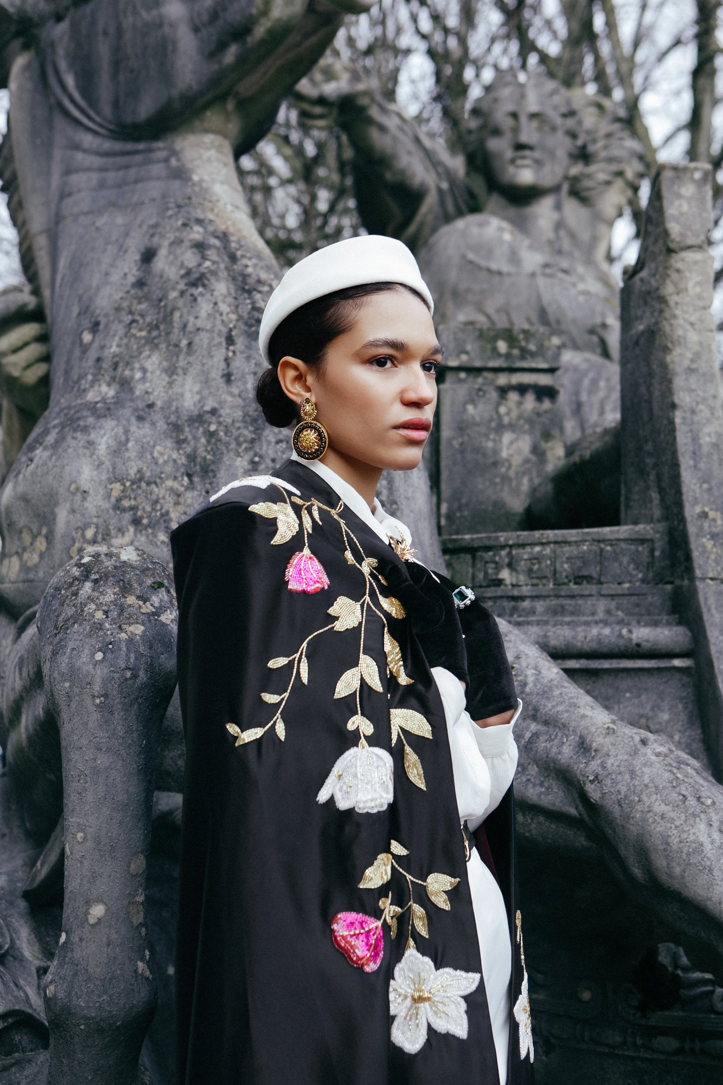 A woman wearing elegant jewelry and a white beret, dressed in a black embroidered coat with floral details, standing outdoors beside a large stone sculpture of a seated woman with an elaborate hairstyle, surrounded by trees.