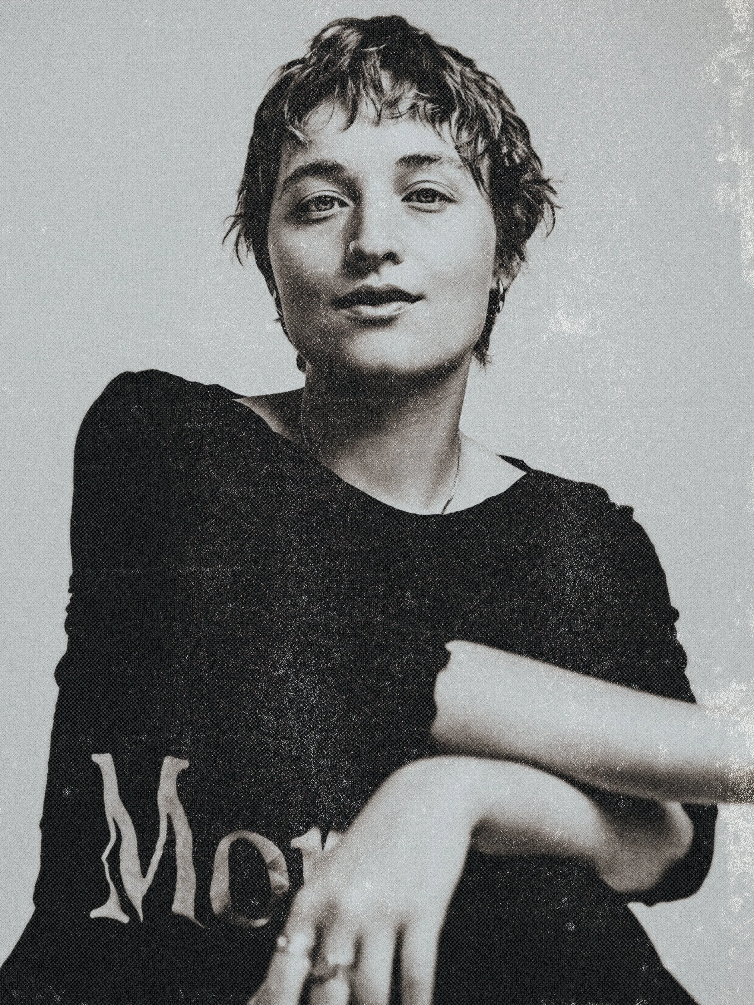 Black and white portrait of a woman with short, wavy hair, wearing a dark top with the word 'MOT' visible on the sleeve, and a necklace. She has a confident expression.