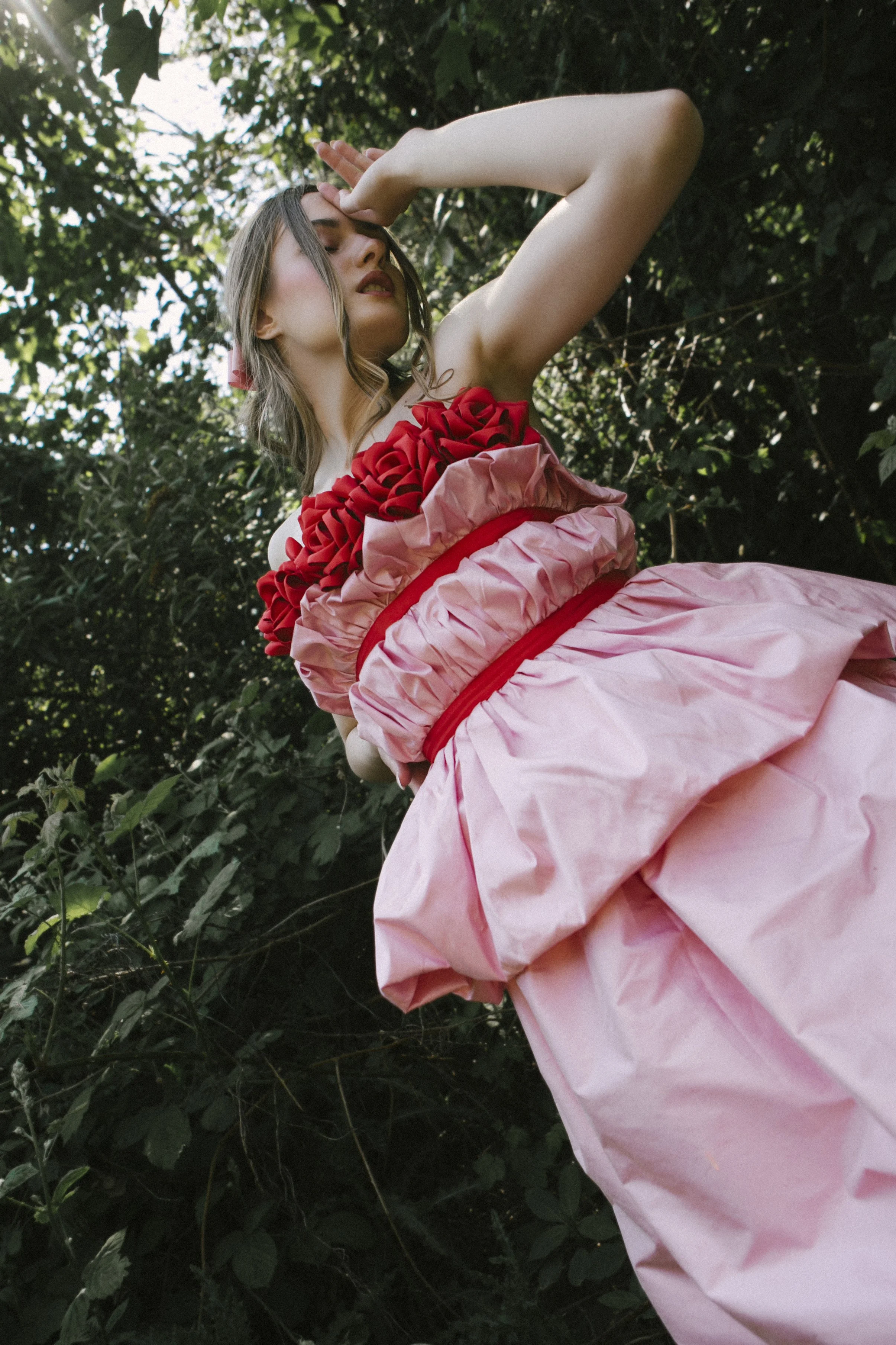 A woman in a pink and red ruffled dress standing outdoors among dense green foliage, with one arm raised and her face partially covered by her hand.