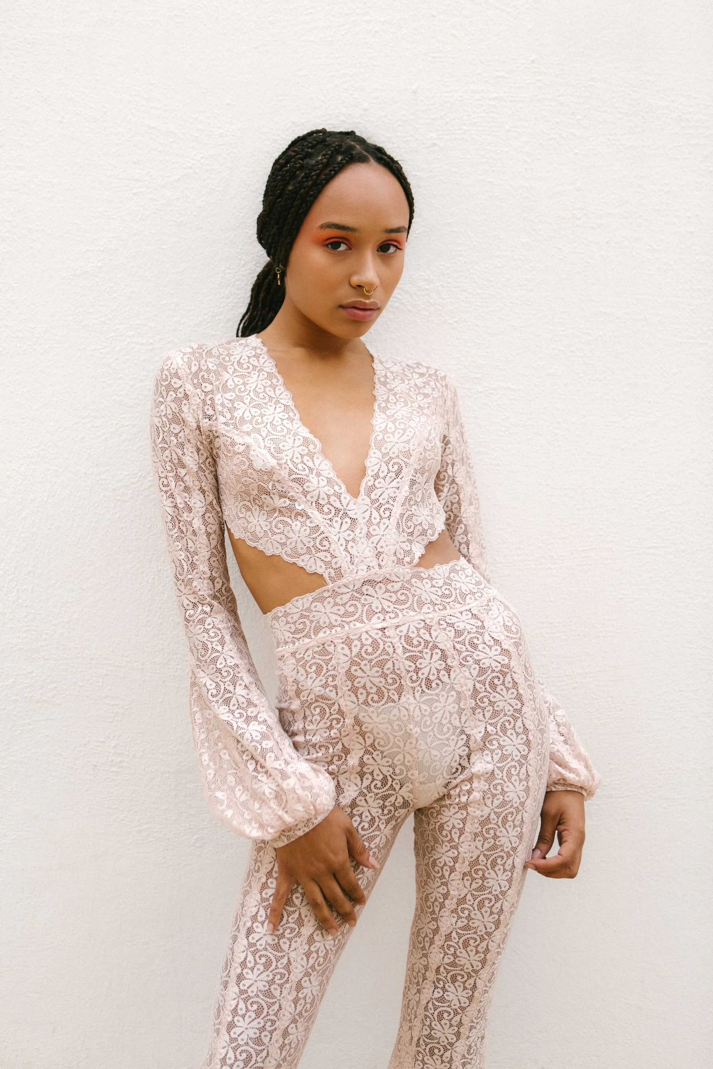 A young woman with dark braided hair wearing a light pink lace crop top and matching pants standing against a plain white wall.