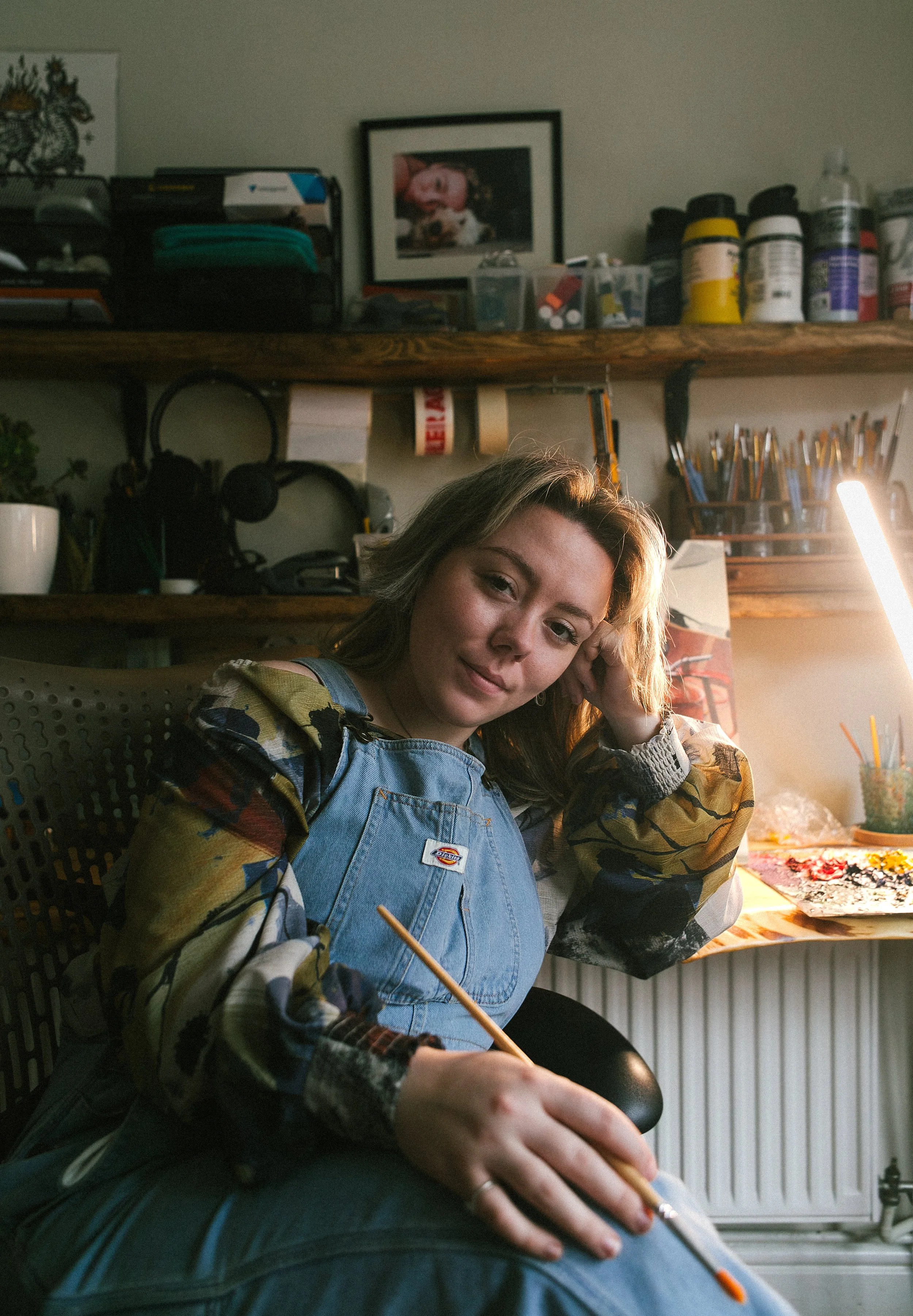 Young woman sitting in an art studio, holding a paintbrush, with art supplies and a palette on the table behind her.