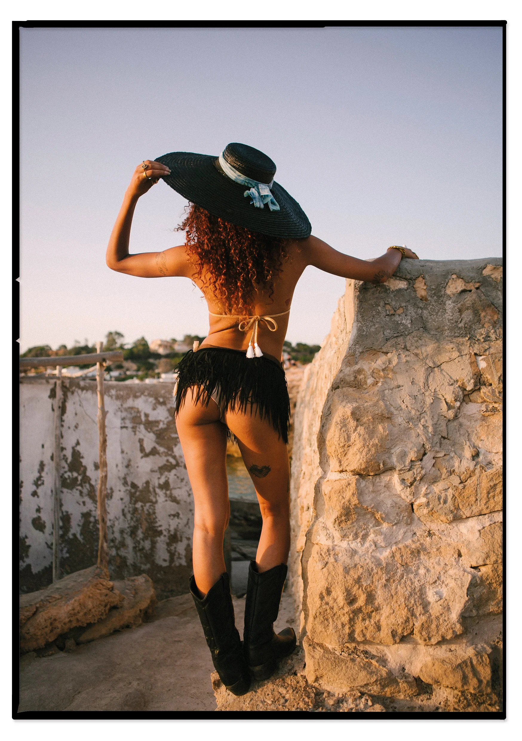 A woman standing outdoors, wearing a large black sun hat with a blue ribbon, a beige bikini top tied at the back, black fringed shorts, and black boots. She is facing away from the camera, holding her hat, with a stone wall and a distant landscape vi
