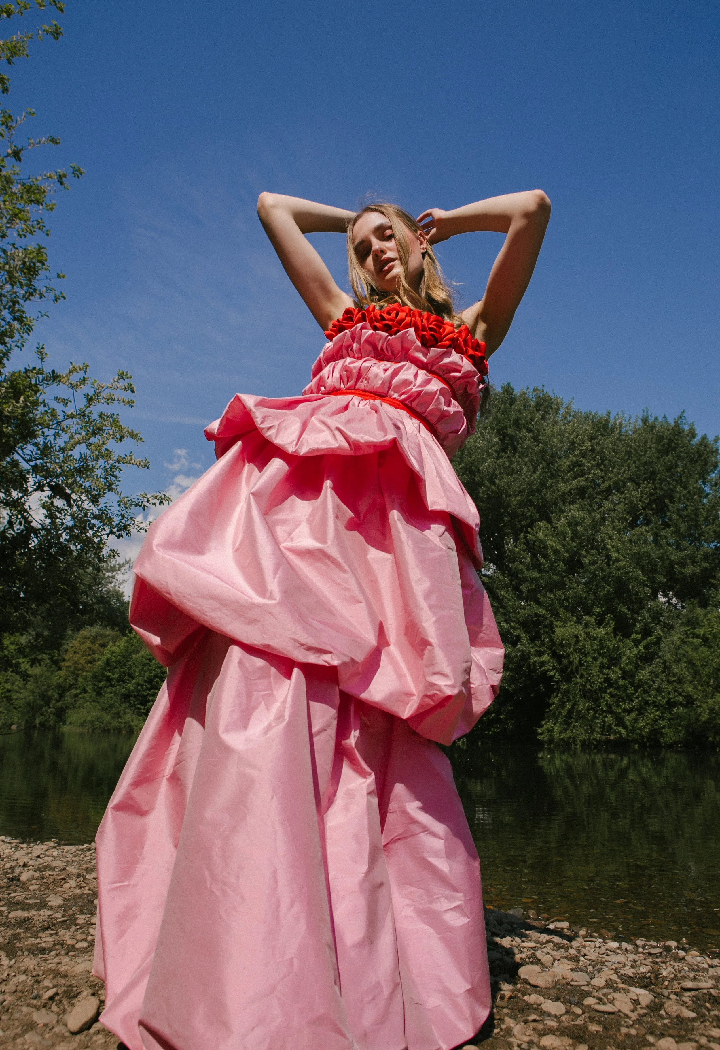 A woman in a pink and red ornate dress standing outdoors near a river with trees, looking down at the camera with her arms raised behind her head against a clear blue sky.