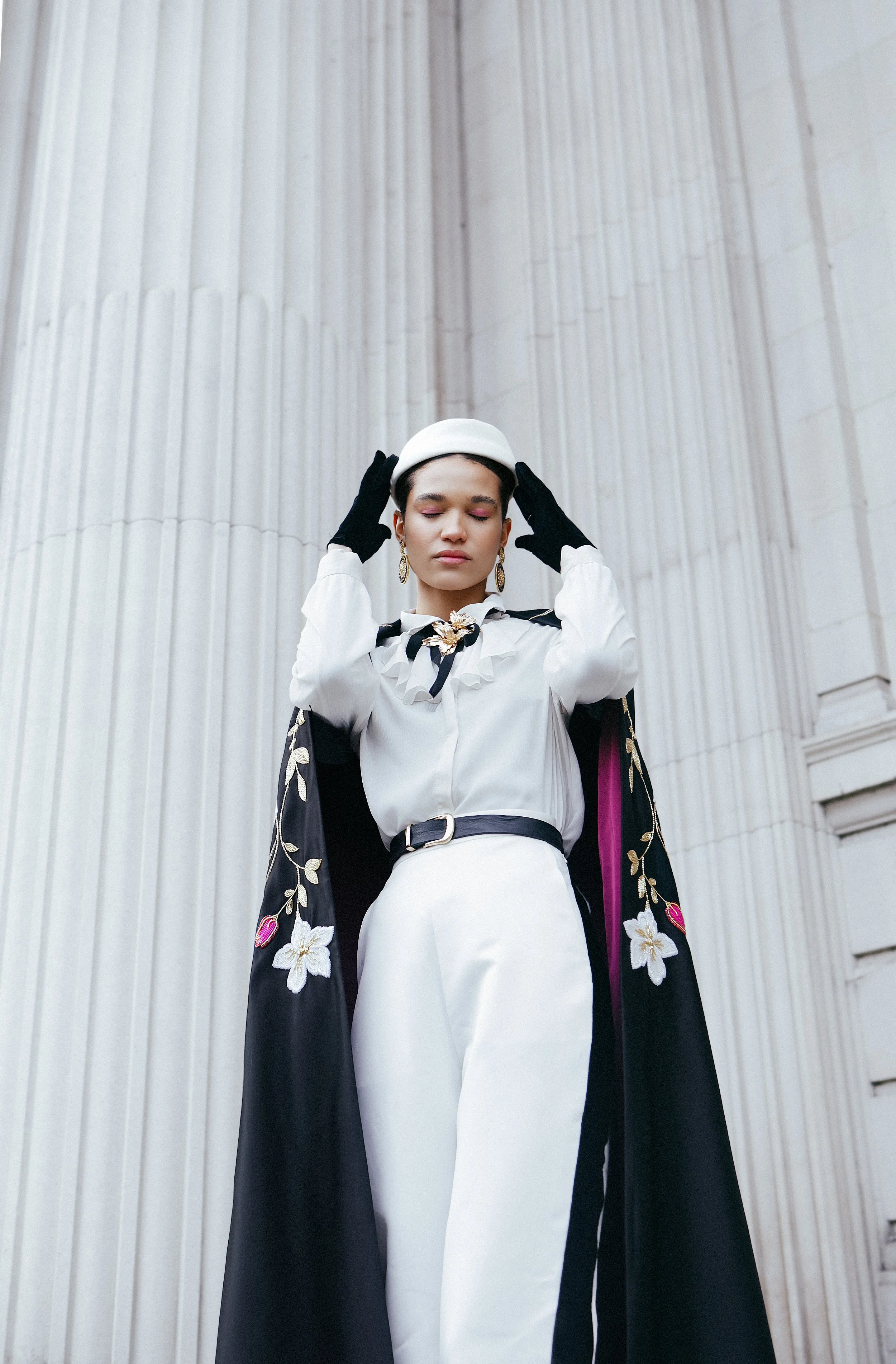 A woman in a white outfit with a black cape featuring floral embroidery, standing in front of large white columns, adjusting a white hat