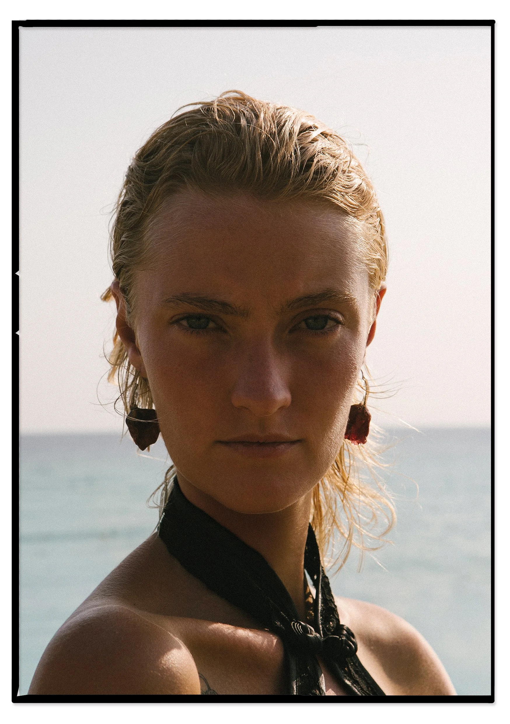 A woman with wet blonde hair standing outdoors near the ocean, looking directly at the camera, with a serious expression. She is wearing earrings and a black scarf or bandana.