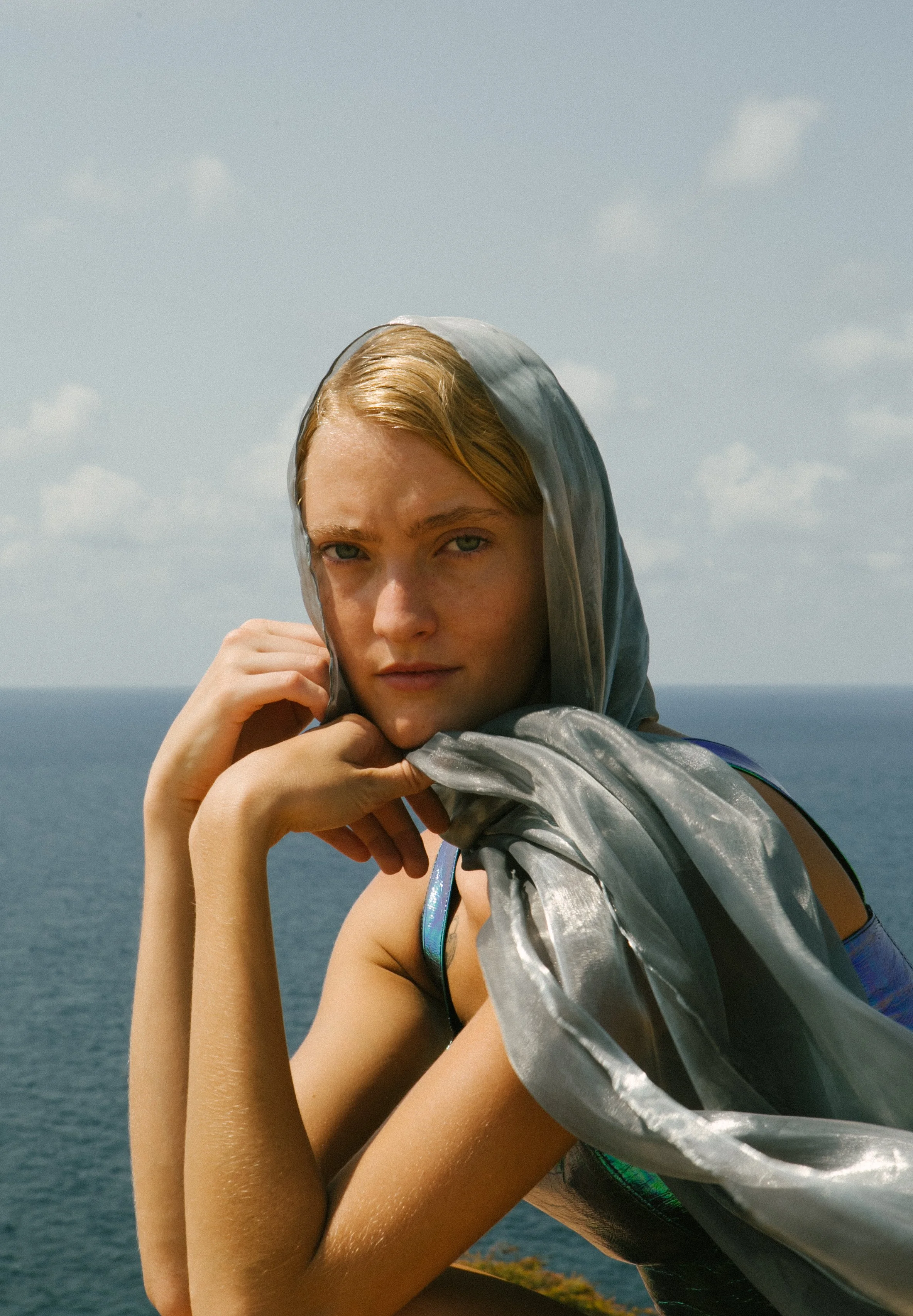A young woman with blonde hair, light skin, and blue eyes, gazing into the camera, with a silver metallic shimmery scarf around her head and shoulders, standing outdoors near the ocean under a partly cloudy sky.