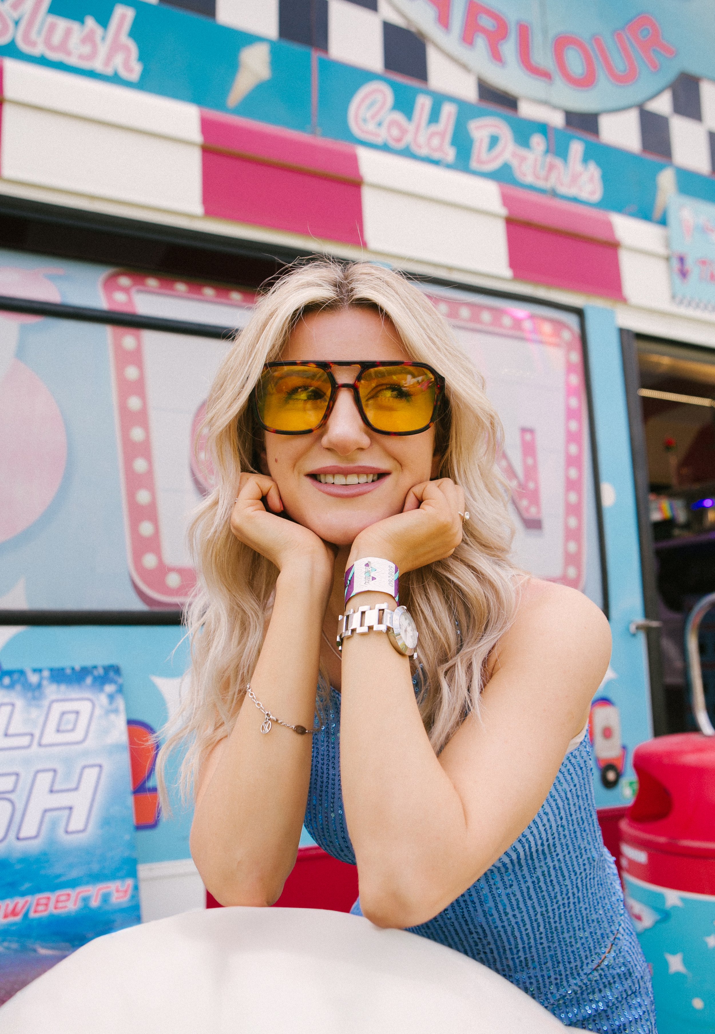 A smiling woman with blond hair and large yellow sunglasses, wearing a blue sleeveless top, sitting outside an ice cream truck with colorful signage and decorations.