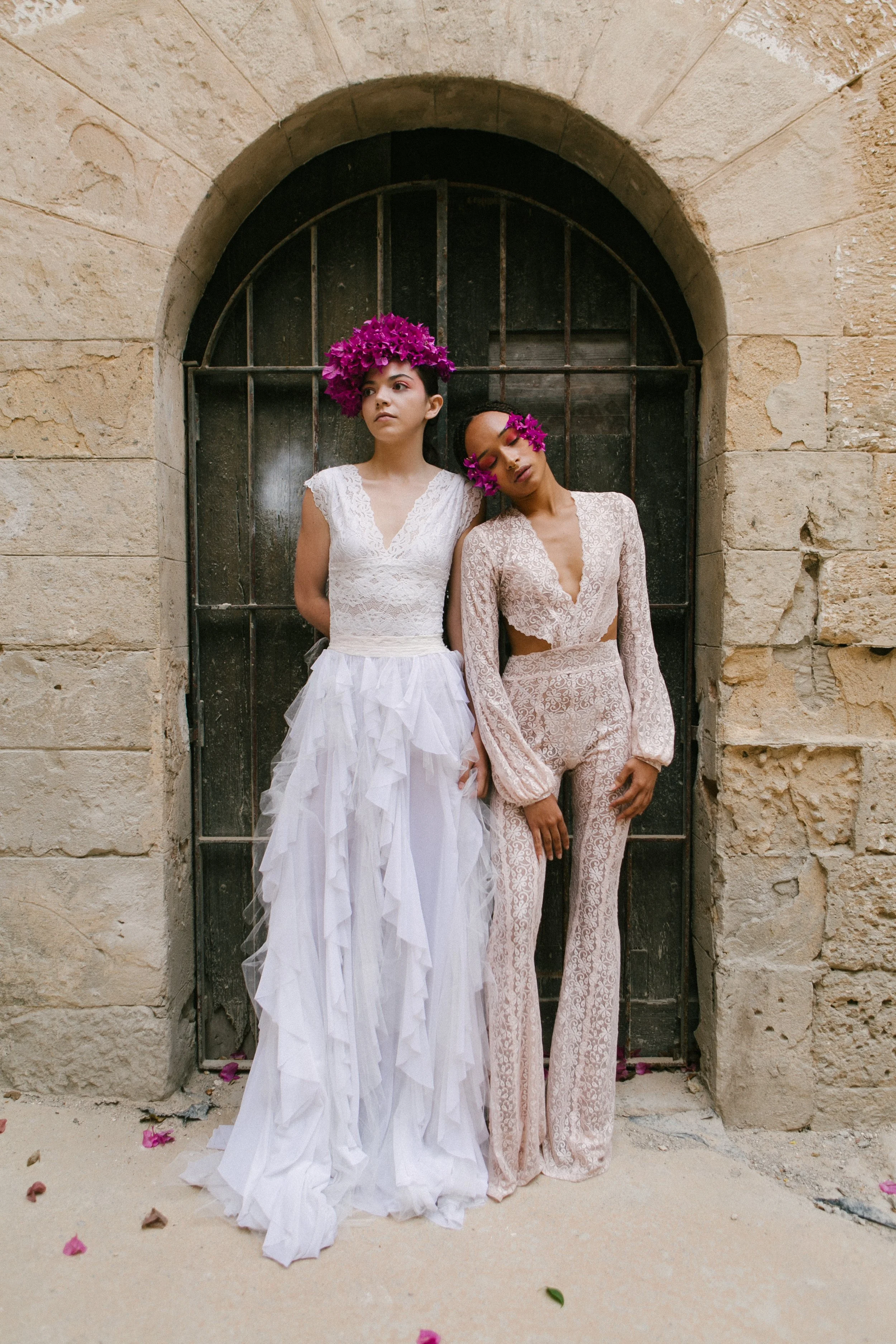 Two women standing in front of a stone wall and black iron gate, wearing white and beige lace outfits with pink flower headpieces.