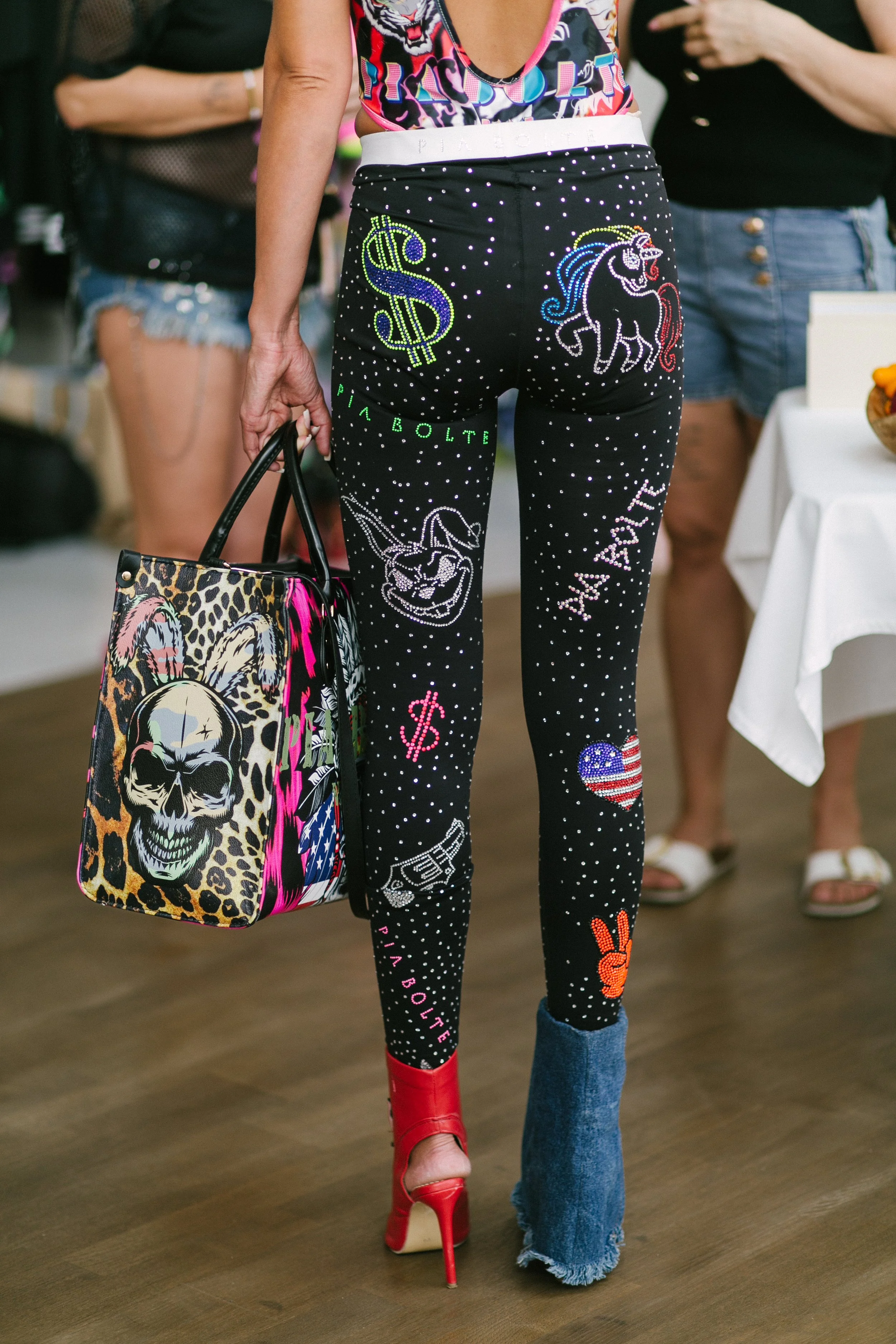 Woman wearing black pants with colorful patches and designs, red high heels, and a colorful top, carrying a large handbag with skull and animal print, at an indoor shopping or event setting.