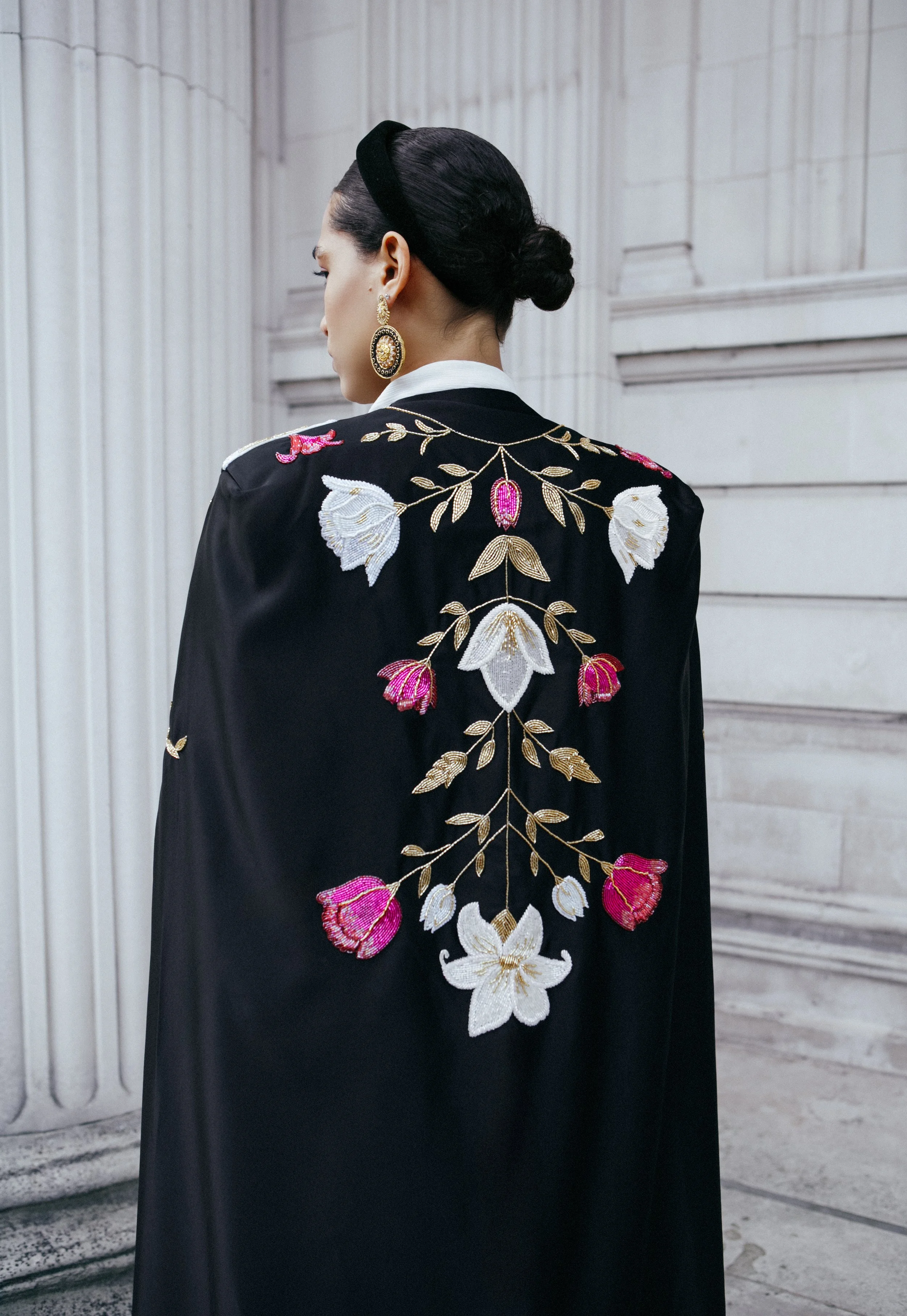 Back view of a woman wearing a black jacket with intricate floral embroidery in white, pink, and gold, standing against a white classical columned background.