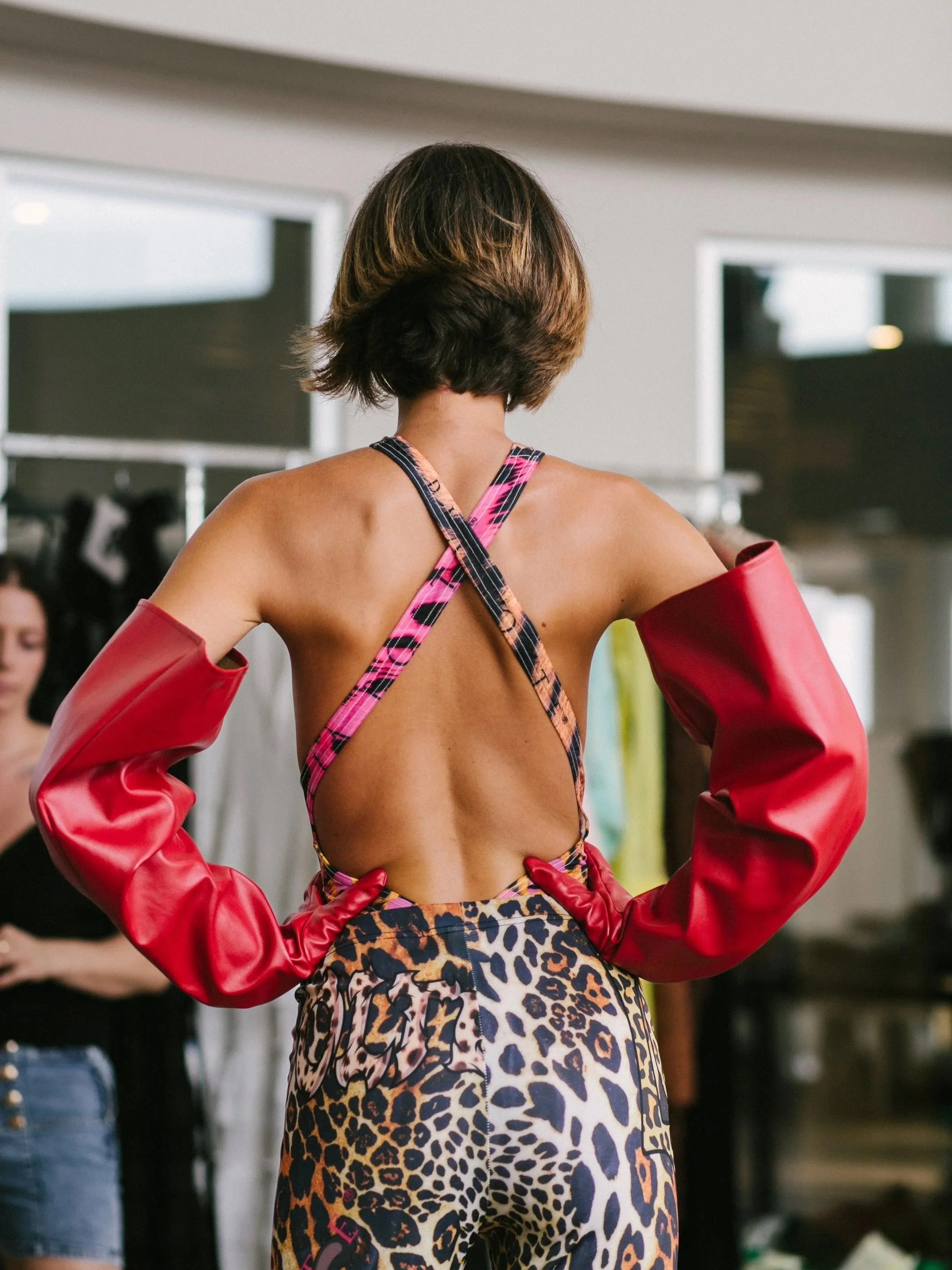 Back view of a woman wearing a colorful animal print crop top with straps and red gloves, standing in a room with other people and clothing in the background.