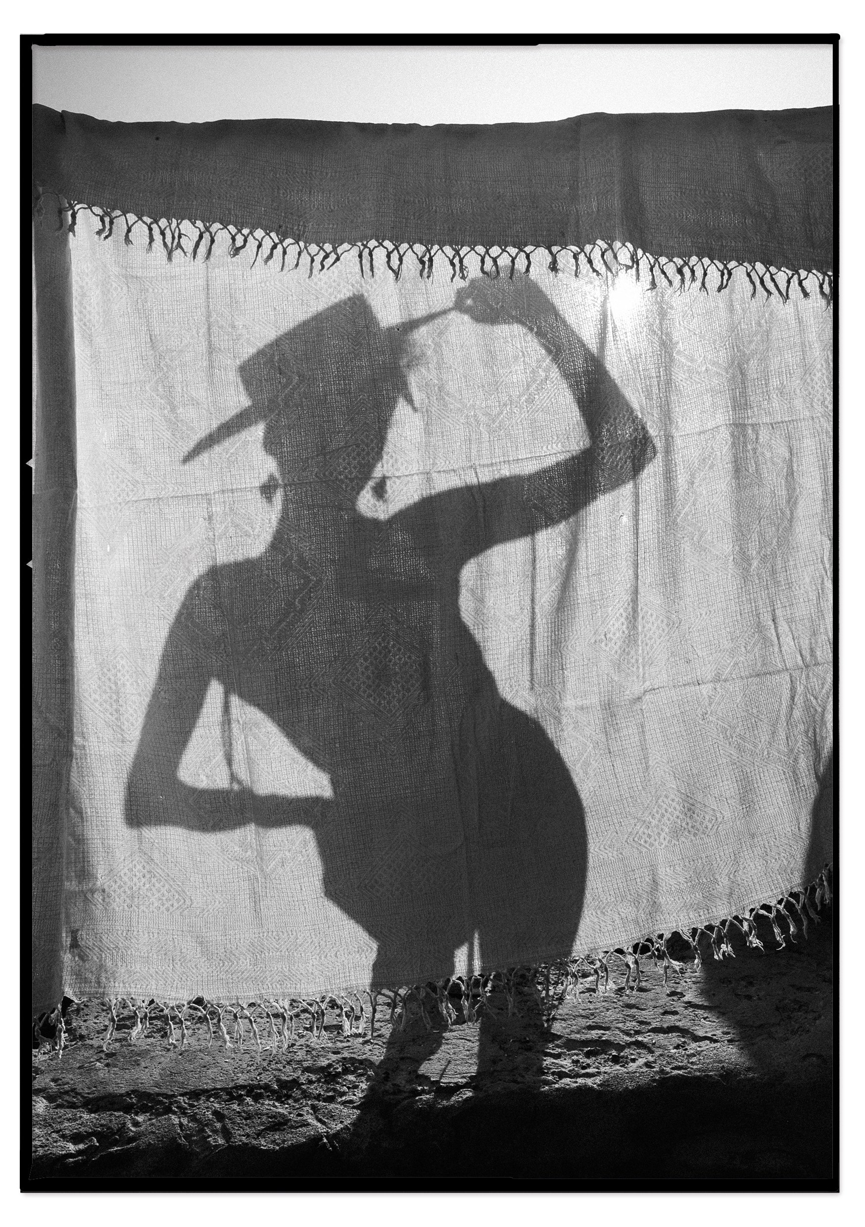 A person’s silhouette is projected onto a fabric curtain, with the sunlight creating a clear shadow. The person is holding a camera or an object to their head, striking a pose, and their shadow emphasizes the figure's stance and arm position.