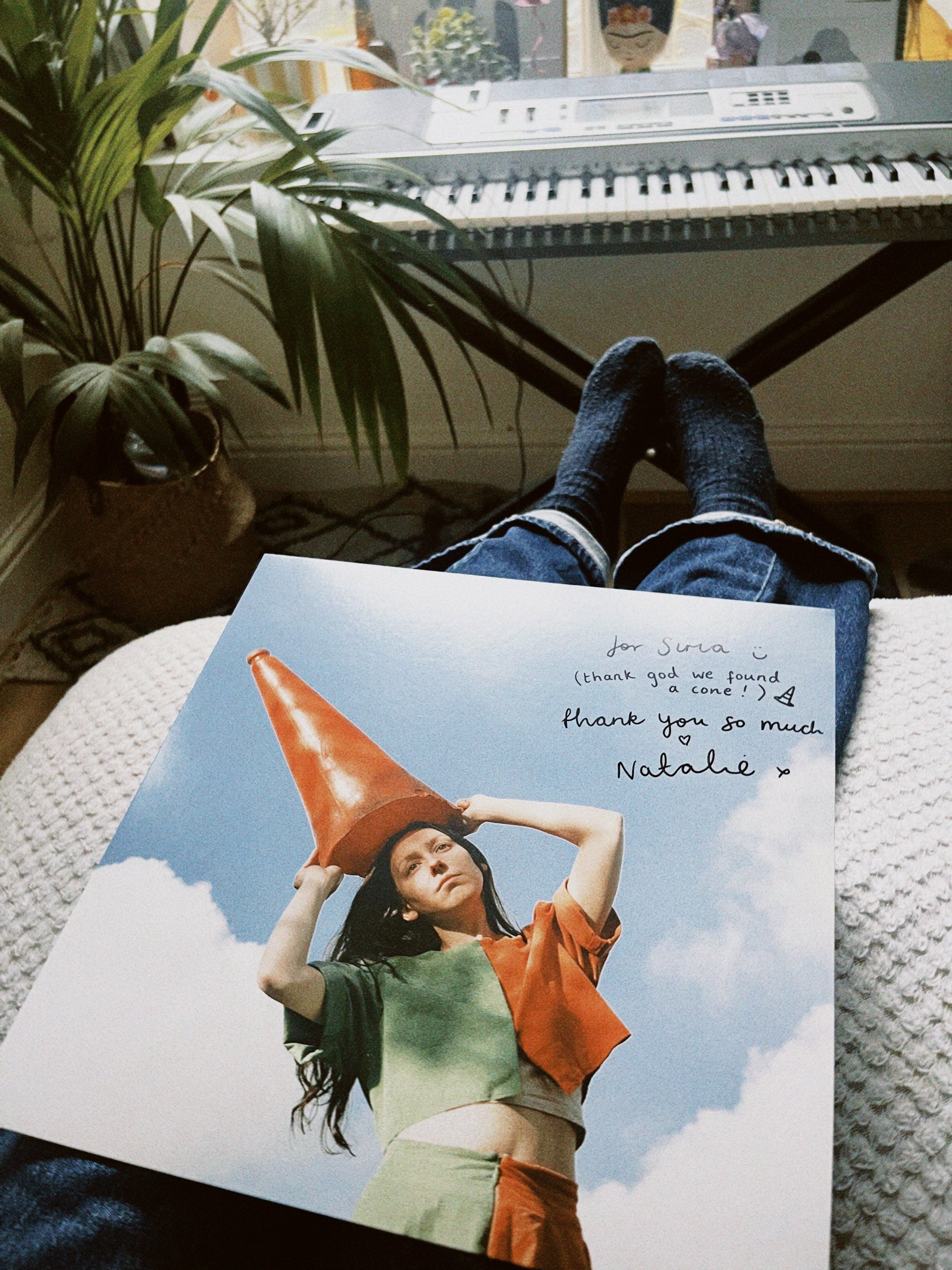 A person sitting on a couch with their legs extended, wearing dark socks and jeans. In front of them is a magazine featuring a woman in a colorful outfit and large orange cone hat, with handwritten notes and a message.