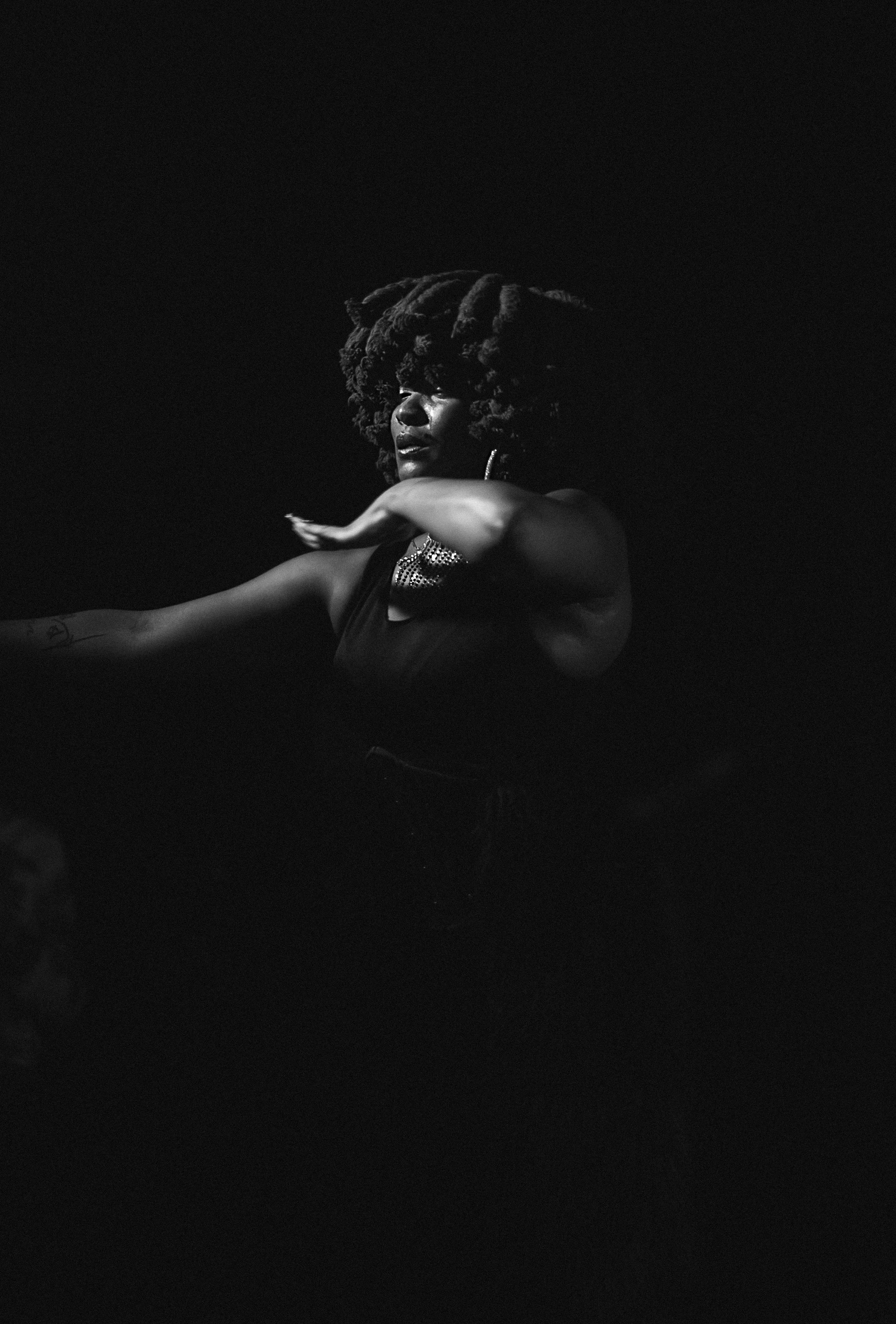A black-and-white photo of a woman with large, curly hair and jewelry, extending her arm forward against a dark background.