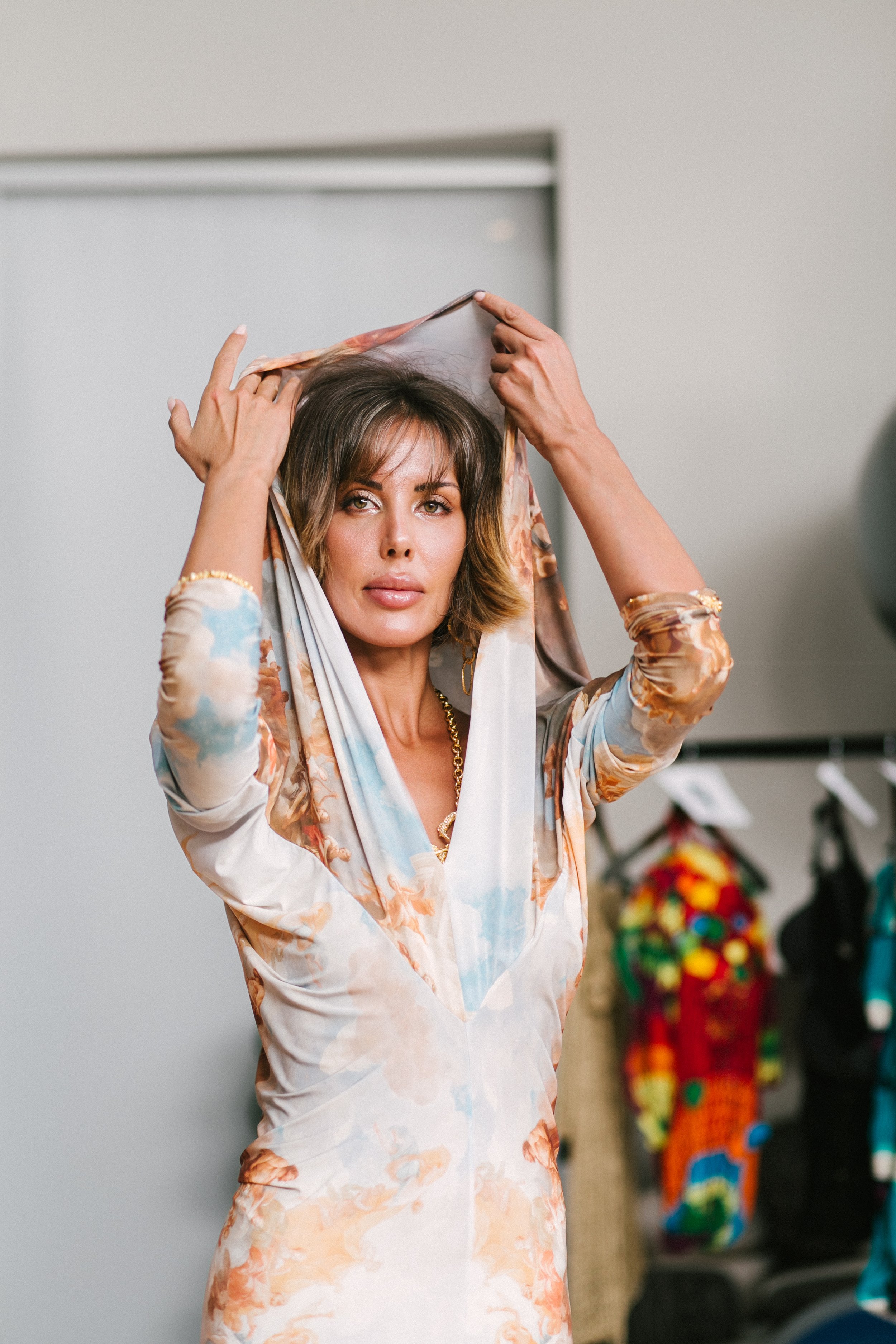 A woman with shoulder-length hair is adjusting a floral scarf on her head in an indoor setting with clothing and accessories in the background.