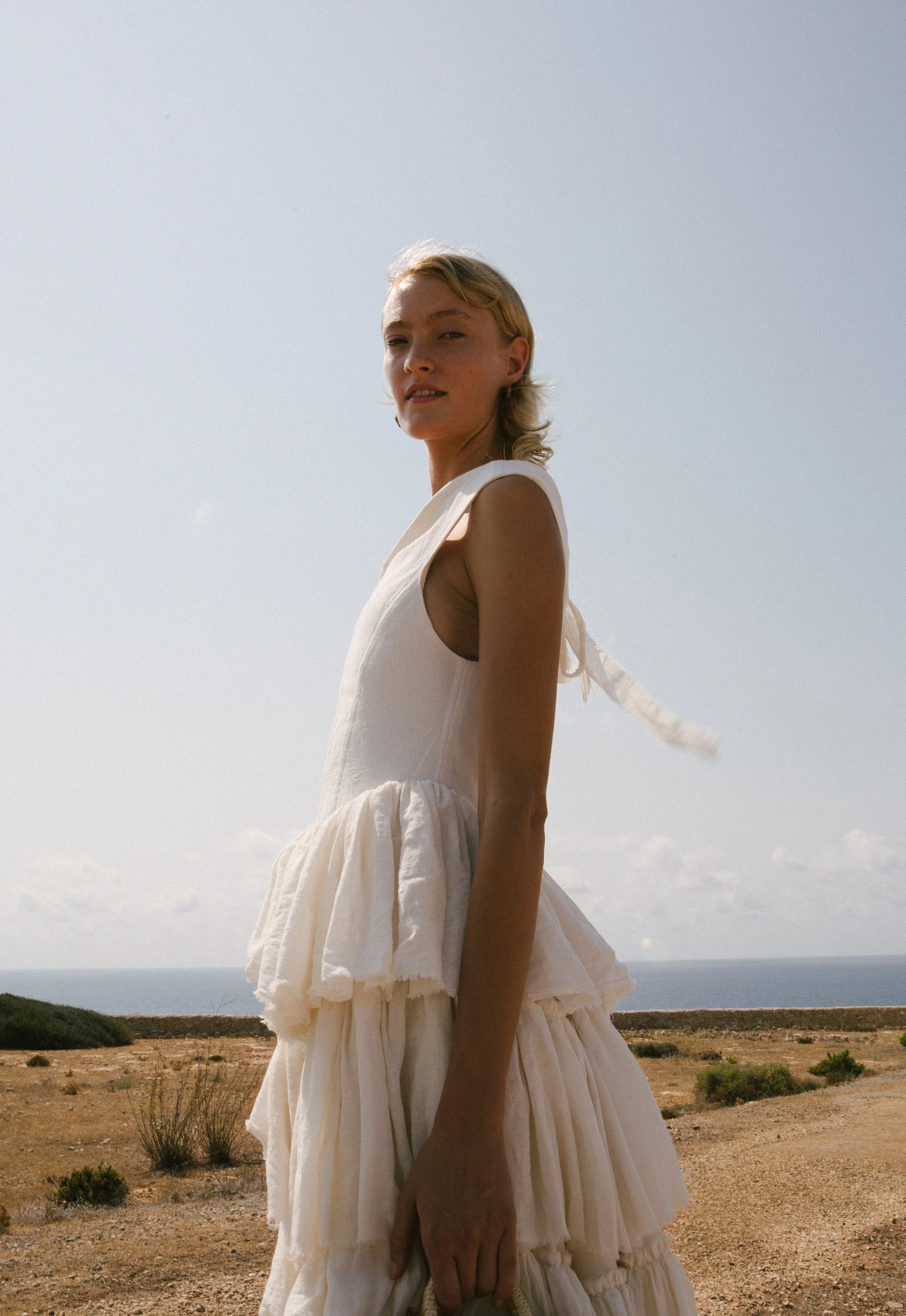 A woman wearing a white dress stands outdoors in a desert-like landscape with a distant view of the ocean and sky in the background.