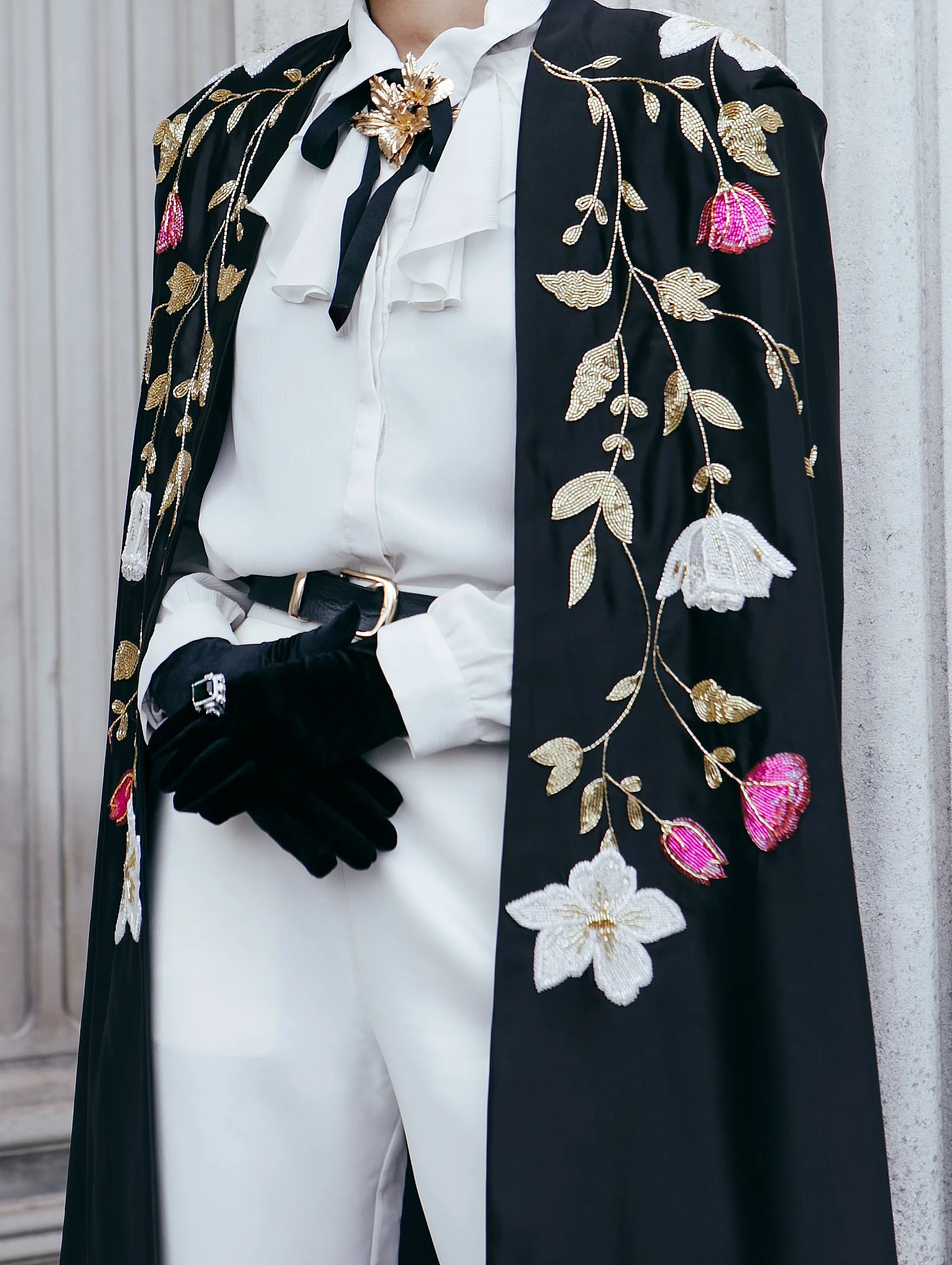 Close-up of a person wearing an embroidered black cape with floral patterns over a white blouse, black gloves, and white pants. The outfit features detailed gold and pink embroidery.