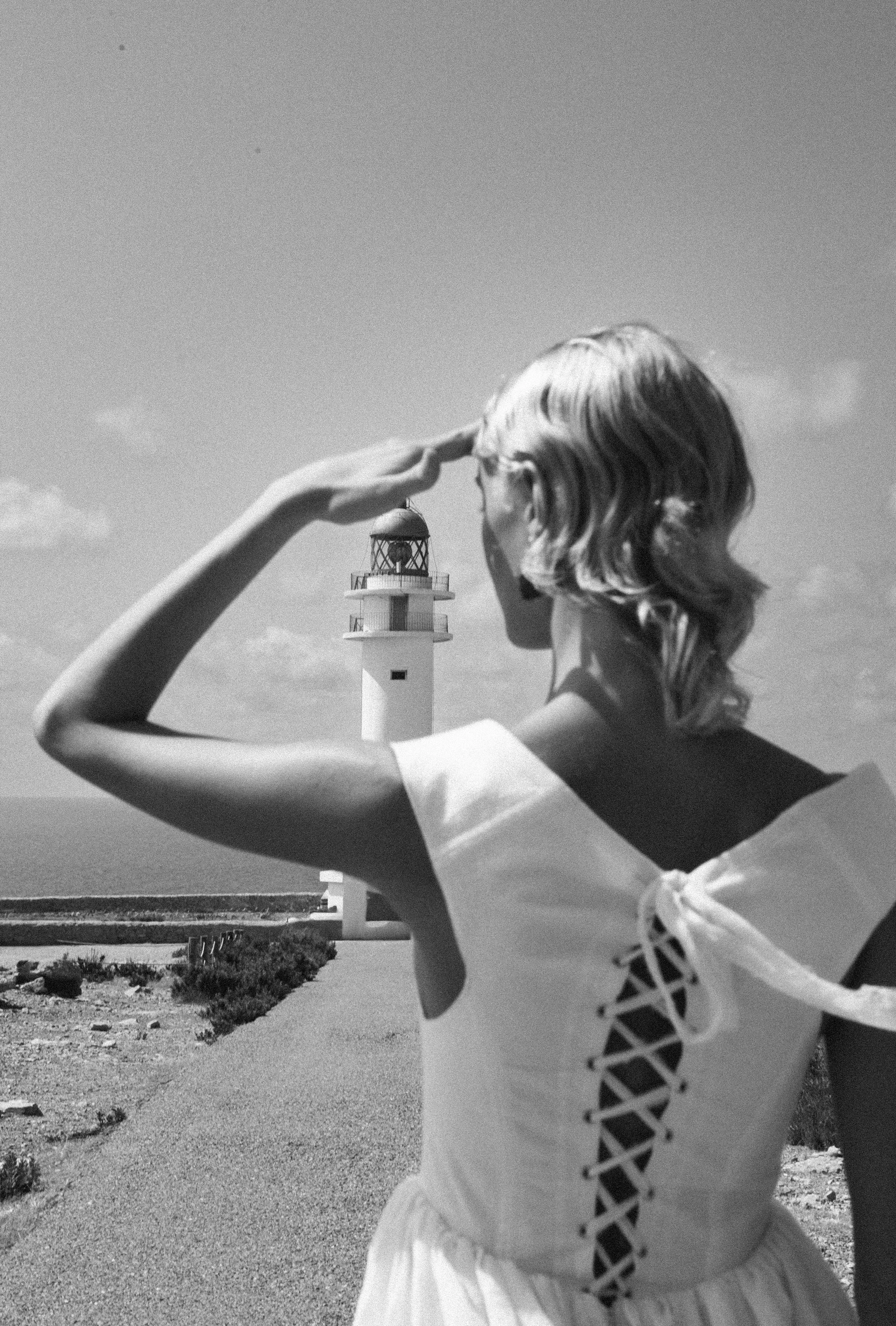 A woman in a lace-up top looks through a telescope with a lighthouse in the background, on a clear day.