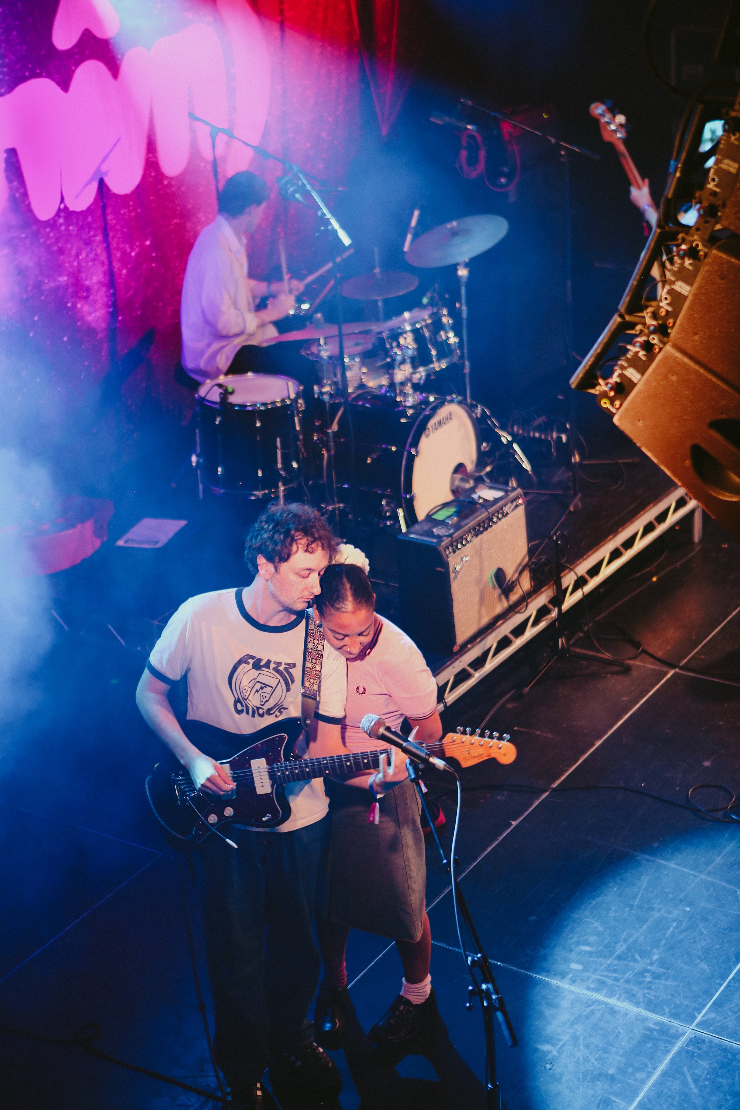 A band performing on stage with a drummer at the back, a guitarist and a singer at the front, lit with colorful stage lights.