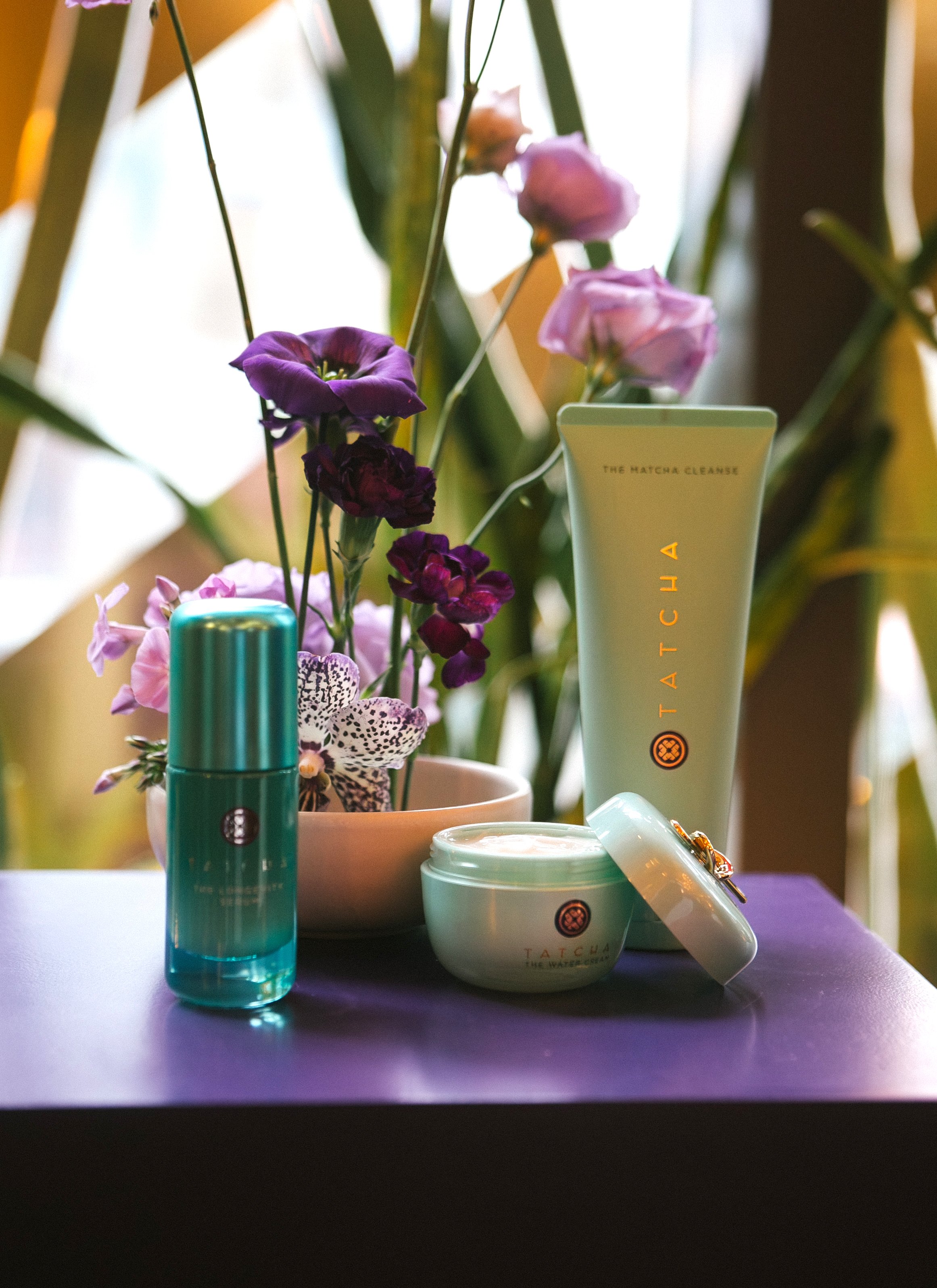 An assortment of skincare products from Tatcha placed on a purple surface, with a plant featuring purple and white flowers in the background.
