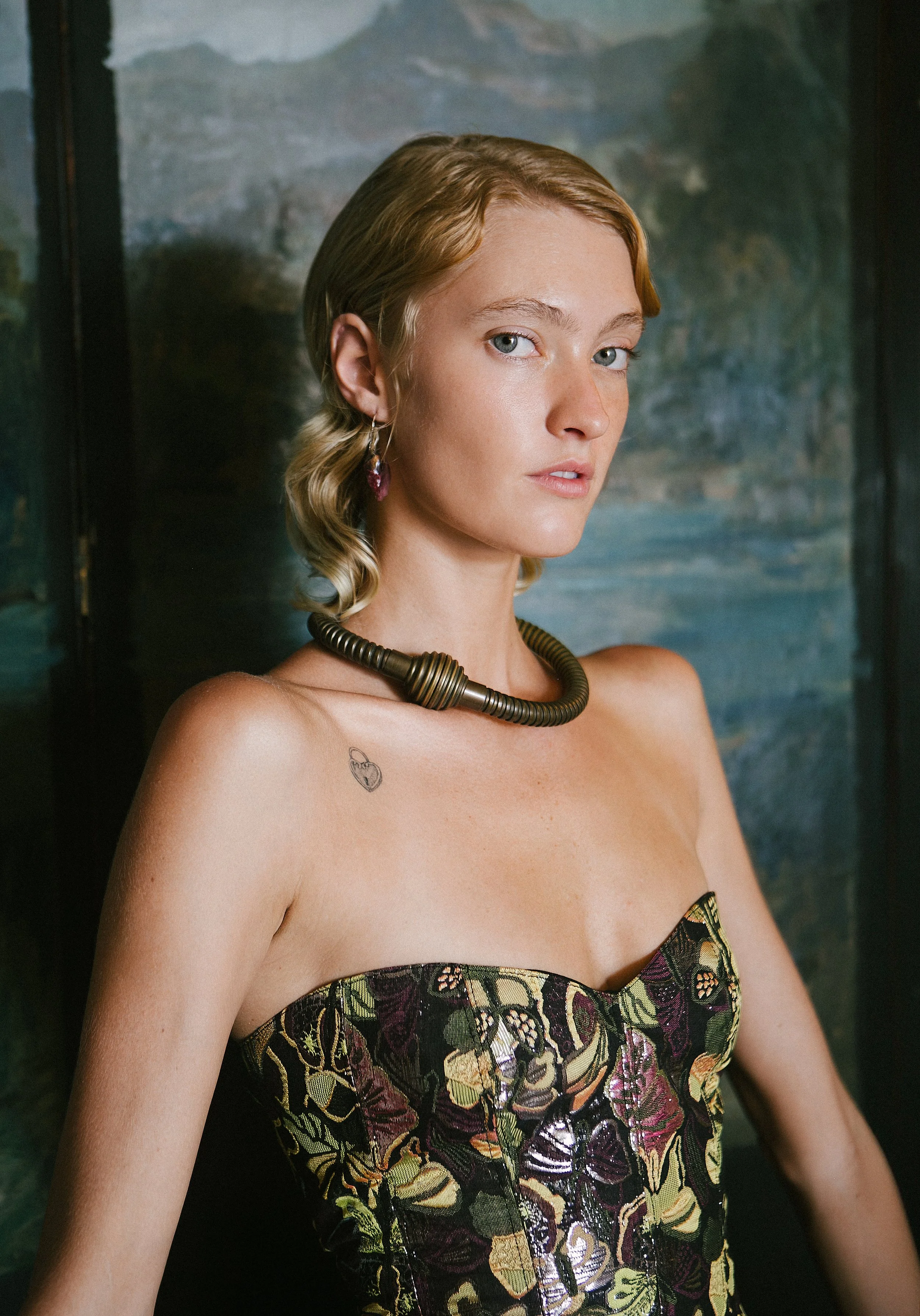 A woman with short, wavy blonde hair and blue eyes posing against a dark, textured background. She is wearing a strapless dress with a butterfly pattern, a chunky metallic necklace, and a dangling earring. She has a small tattoo on her left shoulder.