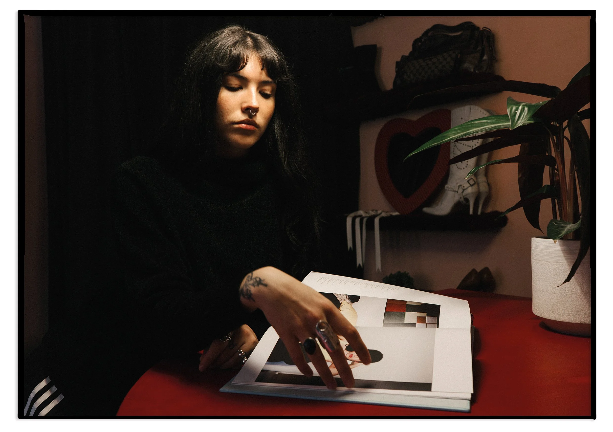 Young woman with black hair and tattoos reading a magazine at a red table, surrounded by houseplants and decorative items in a dimly lit room.