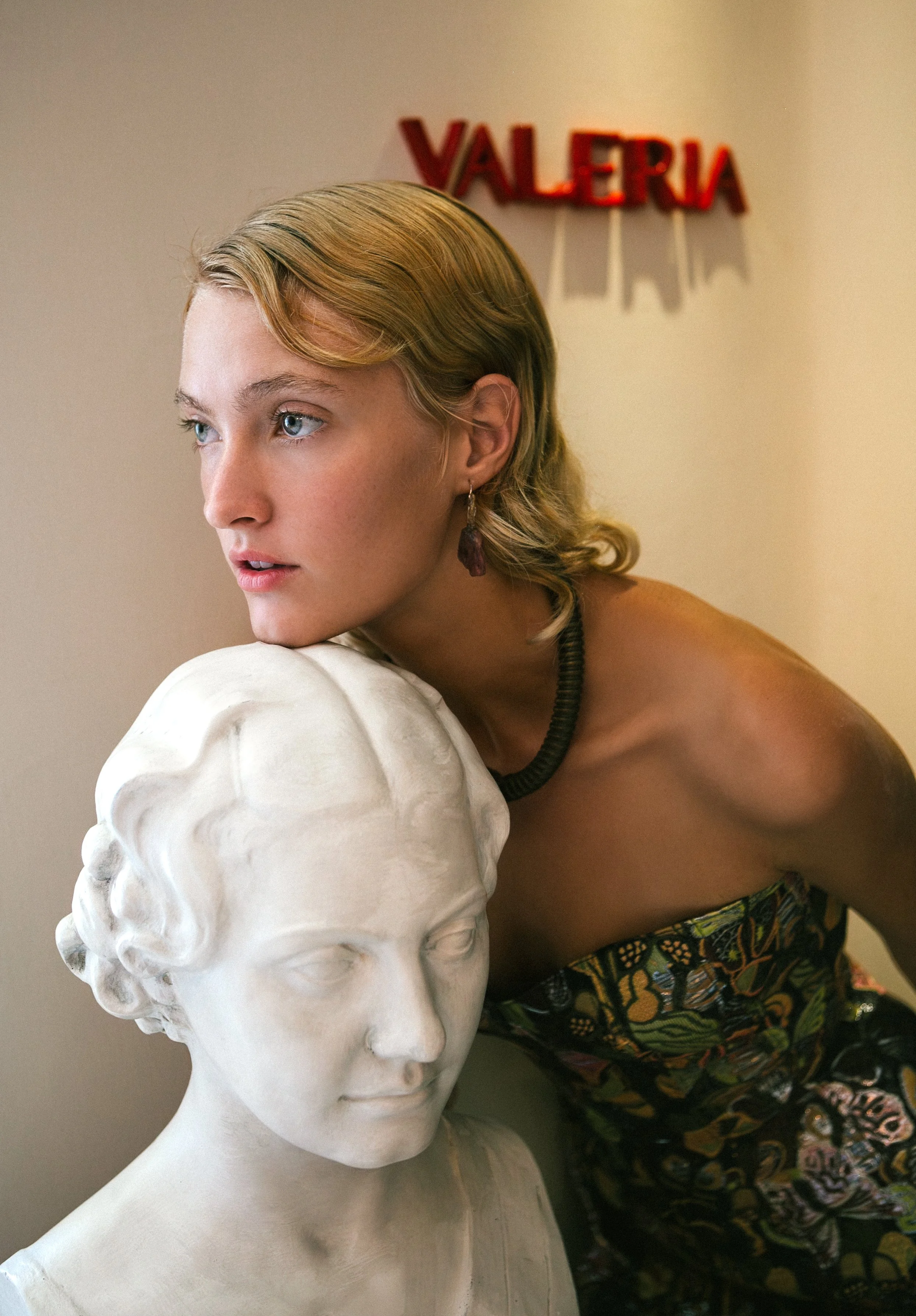 A woman with blonde hair and blue eyes leans on a white bust sculpture of a woman's head. The woman is wearing a floral dress, a black necklace, and a dangling earring. There is a red sign that says 'VALERIA' in the background.
