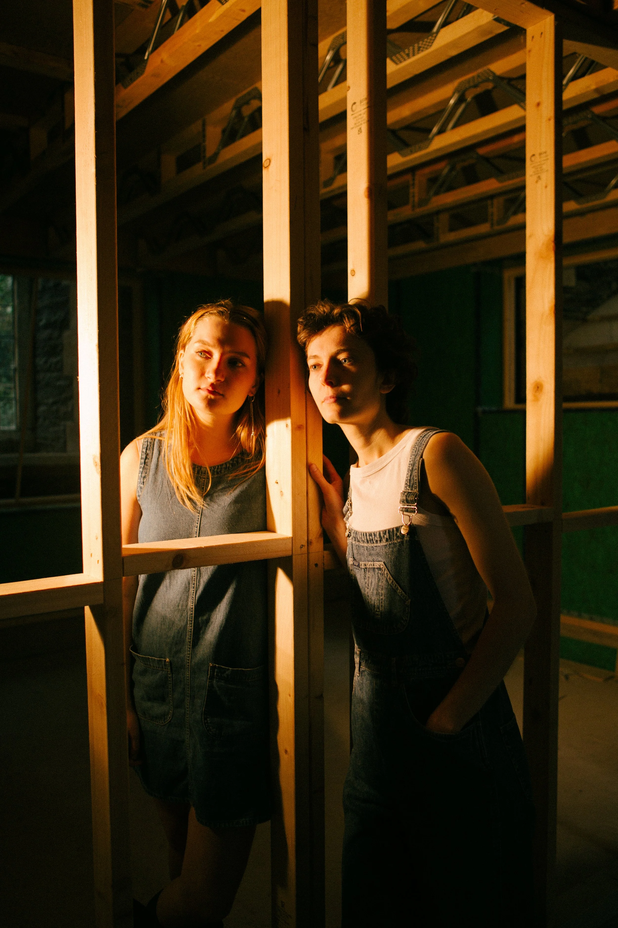 Two young women with denim clothing pose behind a wooden frame structure inside a house under construction, illuminated by warm sunlight.