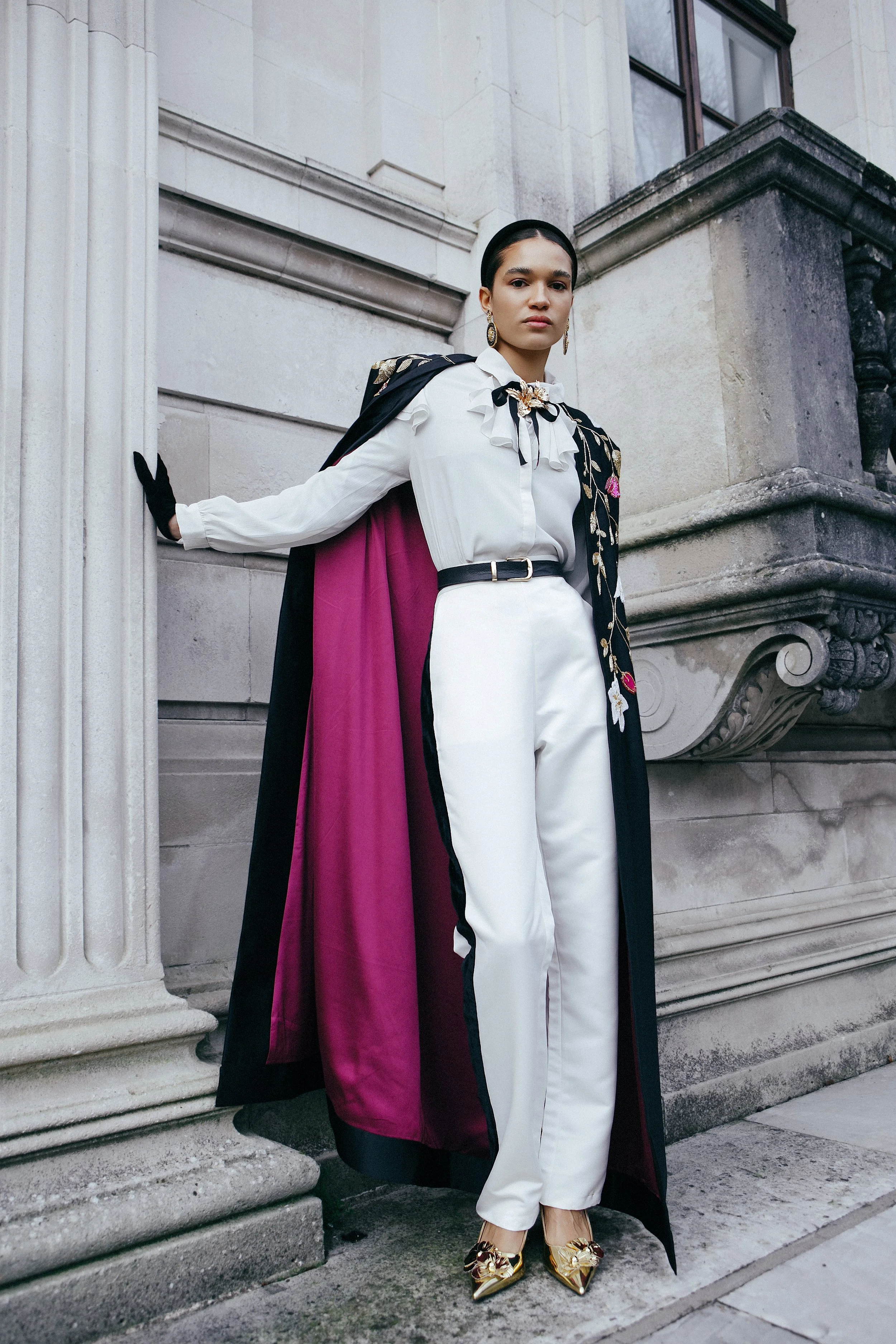 A woman in an elegant white pantsuit with a black belt, gold shoes, and statement jewelry, standing against a stone building with classical architecture.