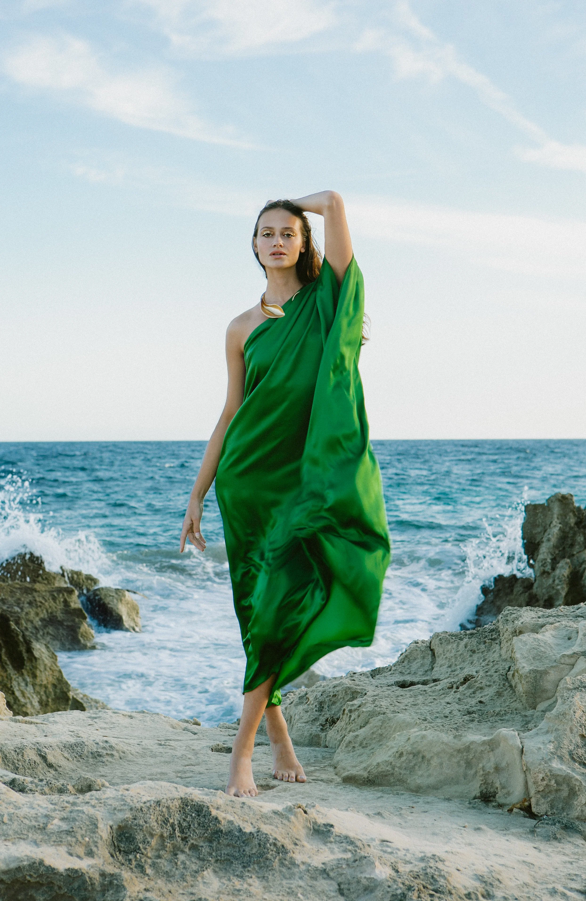 A woman in a green dress standing barefoot on rocks by the ocean, with waves crashing behind her under a partly cloudy sky.