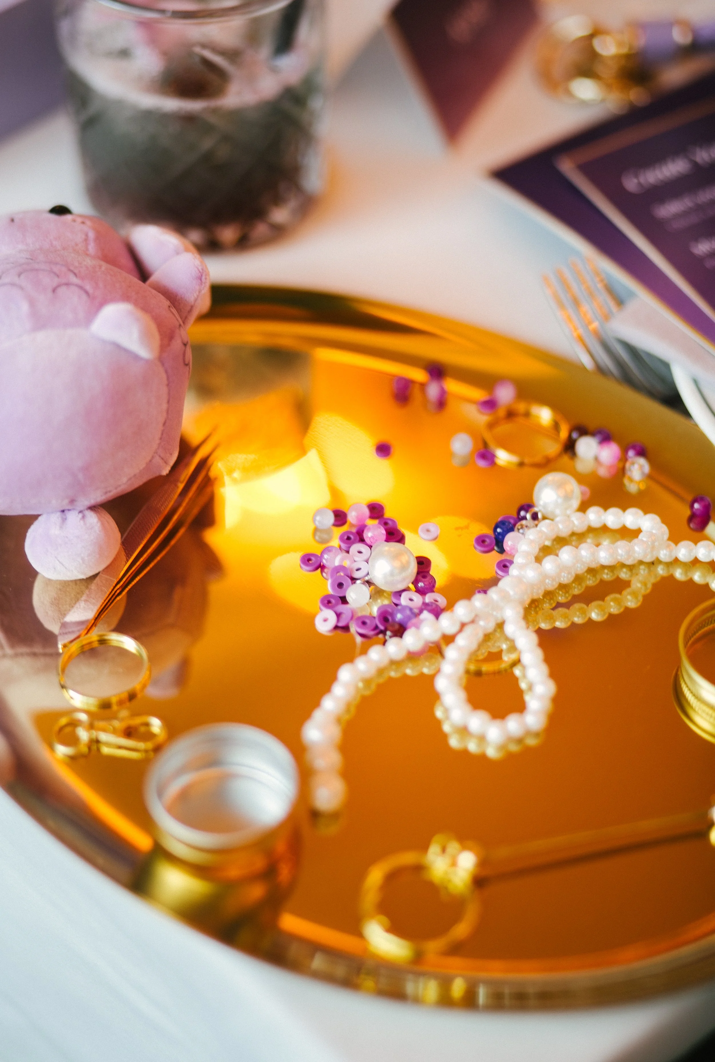 Jewelry and accessories on a reflective gold-colored tray, including pearl necklaces, purple and white sequined earrings, and gold rings, with a pink plush toy on the left and a glass of water and papers in the background.