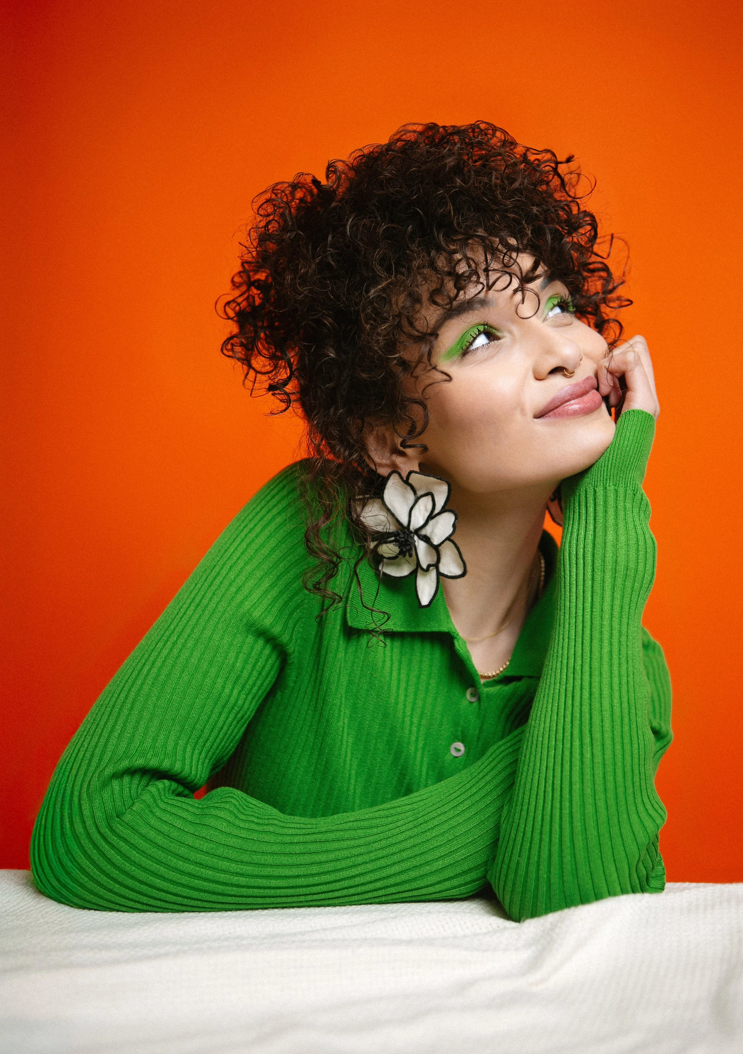 A woman with curly hair and bright green makeup wearing a green ribbed shirt, with a flower-shaped accessory on her ear, resting her chin on her hand against an orange background.