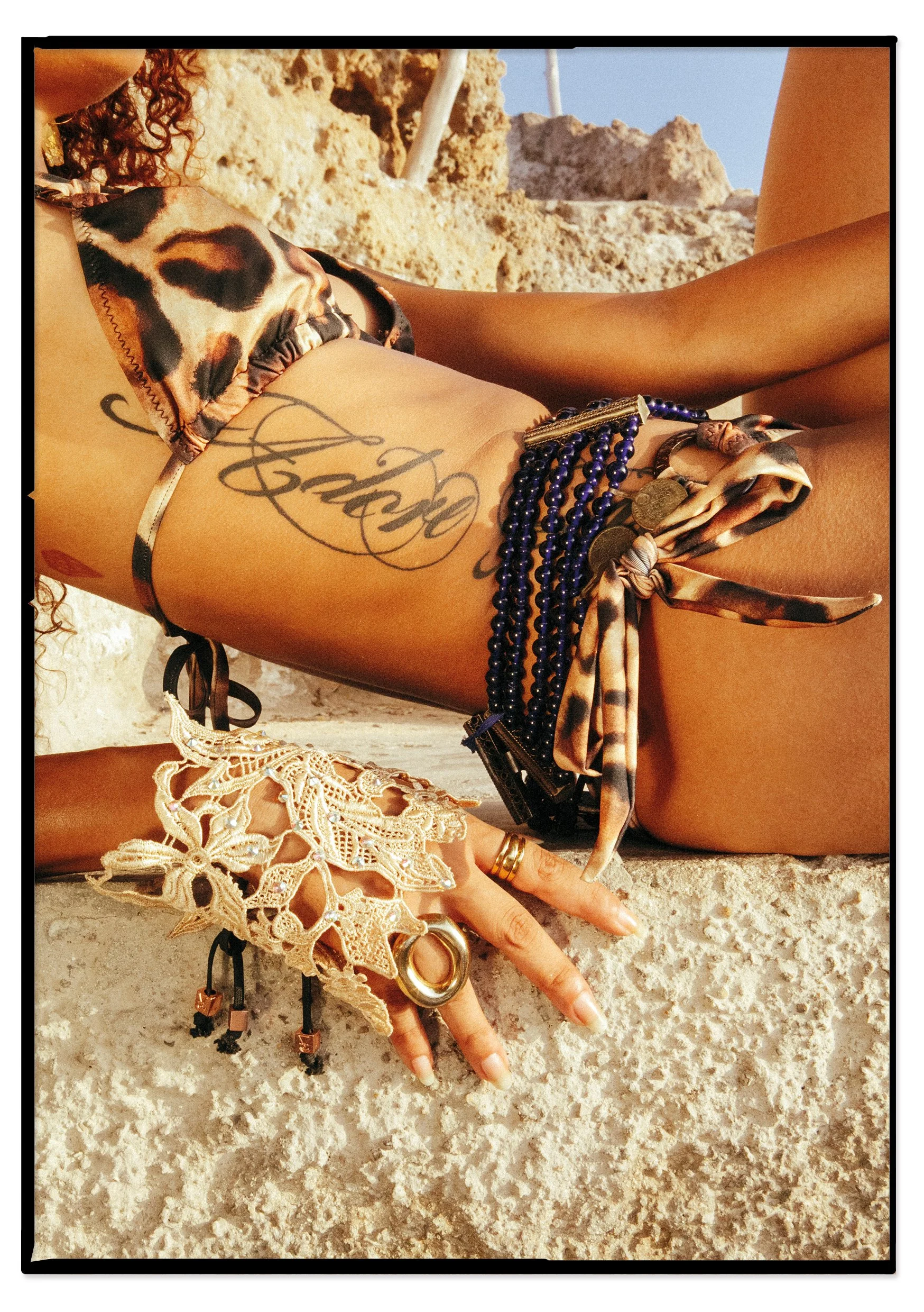 Close-up of a woman's body lying on a rocky surface, showing her tattoos, jewelry, and a swimsuit with animal print. The background features rocks and a clear blue sky.