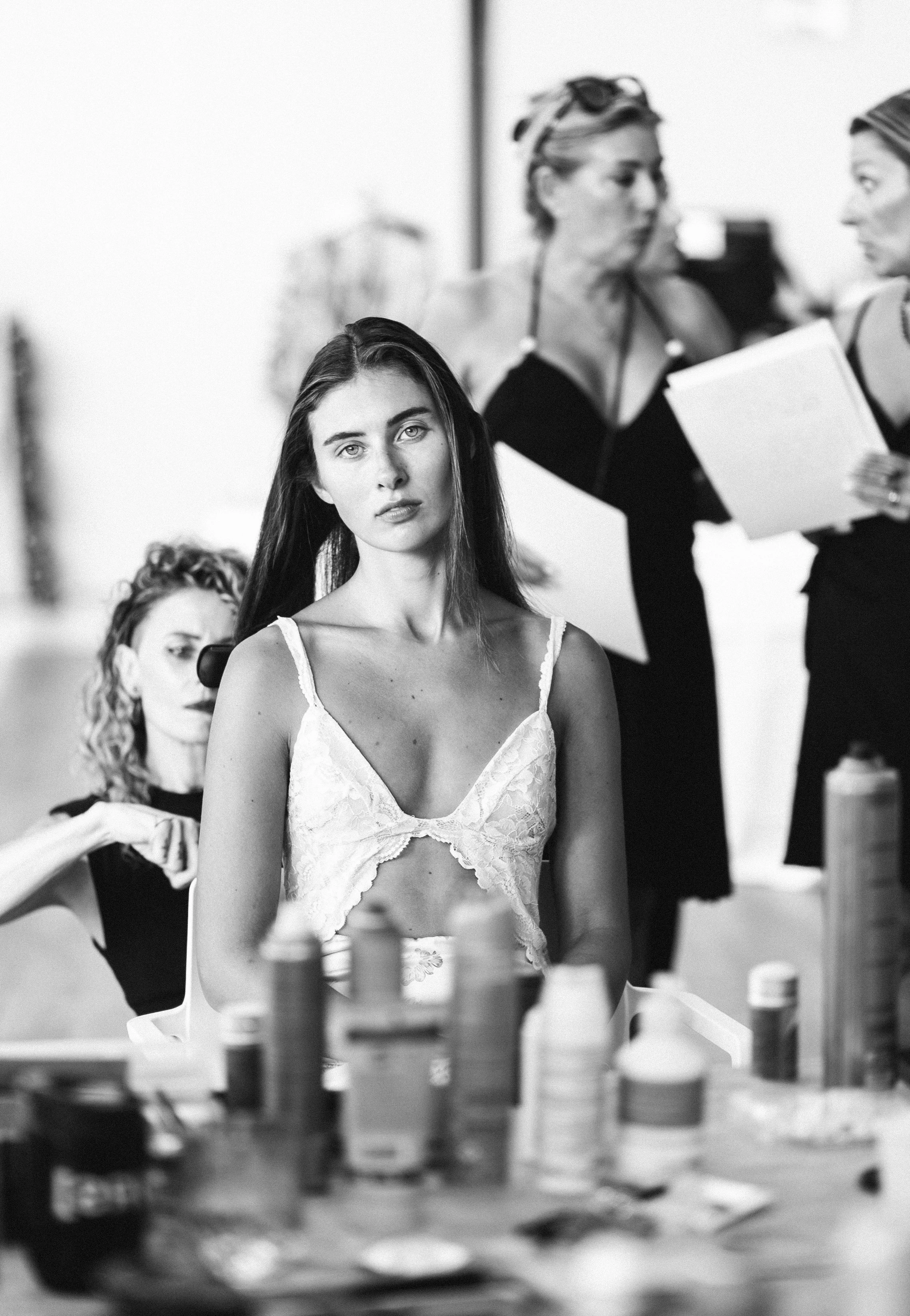 Model in lace top getting makeup done backstage at a fashion show with makeup artists around.