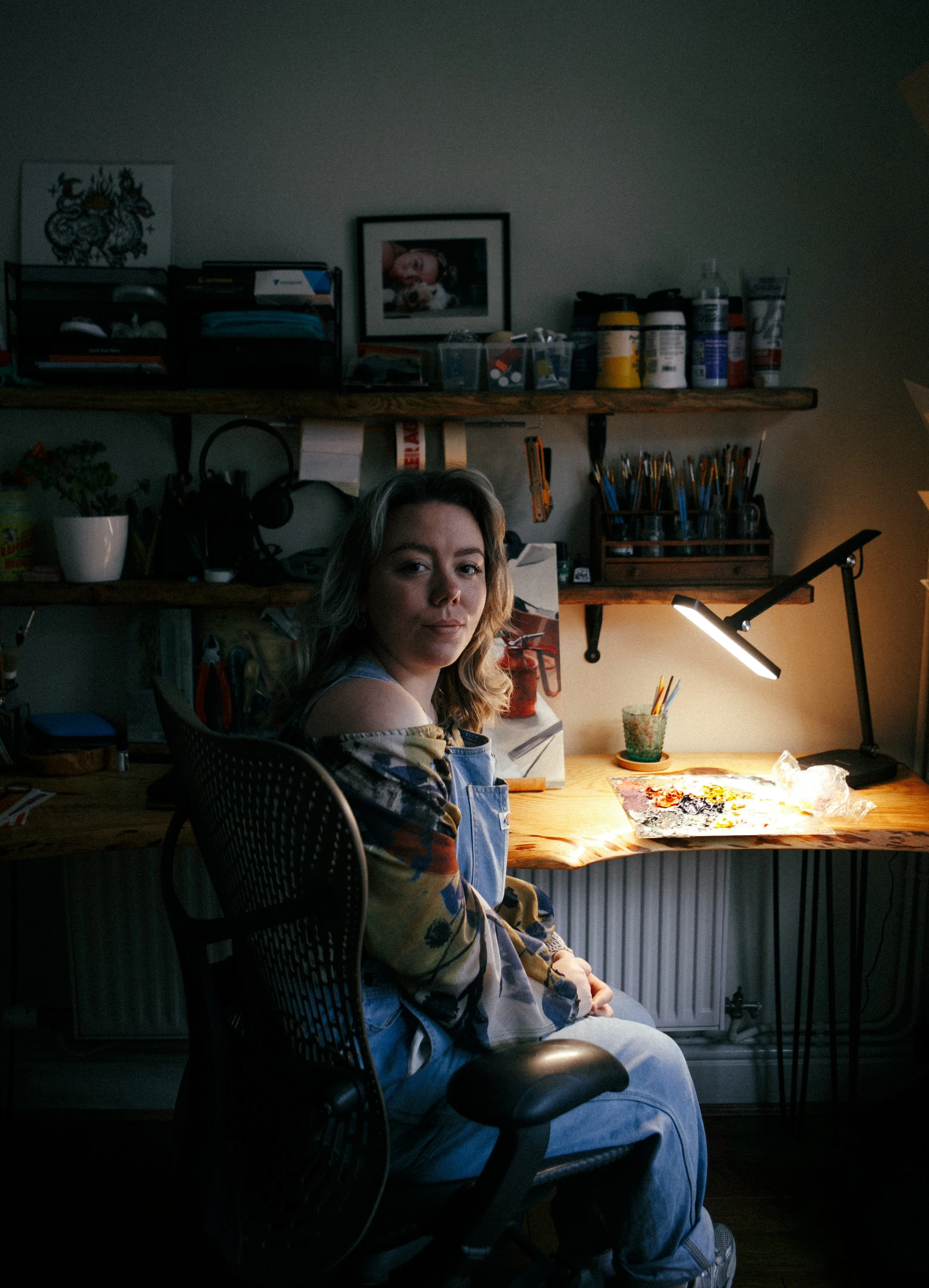 A young woman sitting at a wooden desk in a dimly lit artist's studio, illuminated by a desk lamp, with art supplies on the desk and shelves in the background.