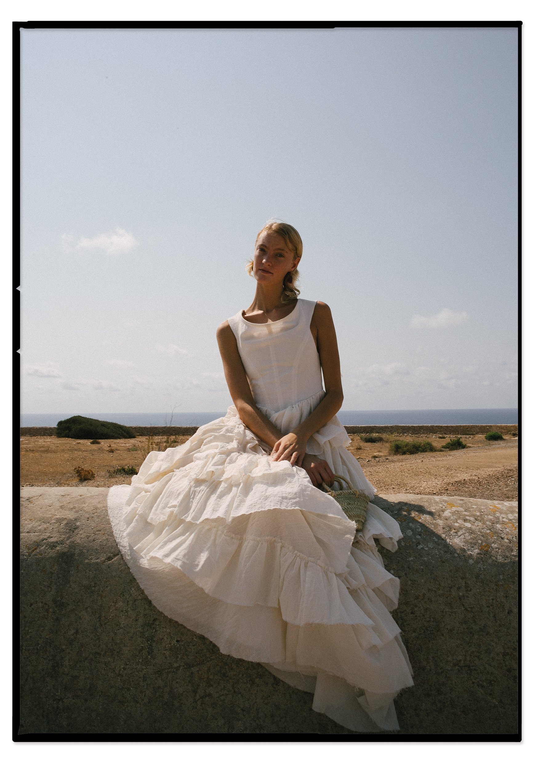 A woman wearing a long, white flowing dress sitting on a large rock outdoors with a flat landscape, some bushes, and a partly cloudy sky in the background.