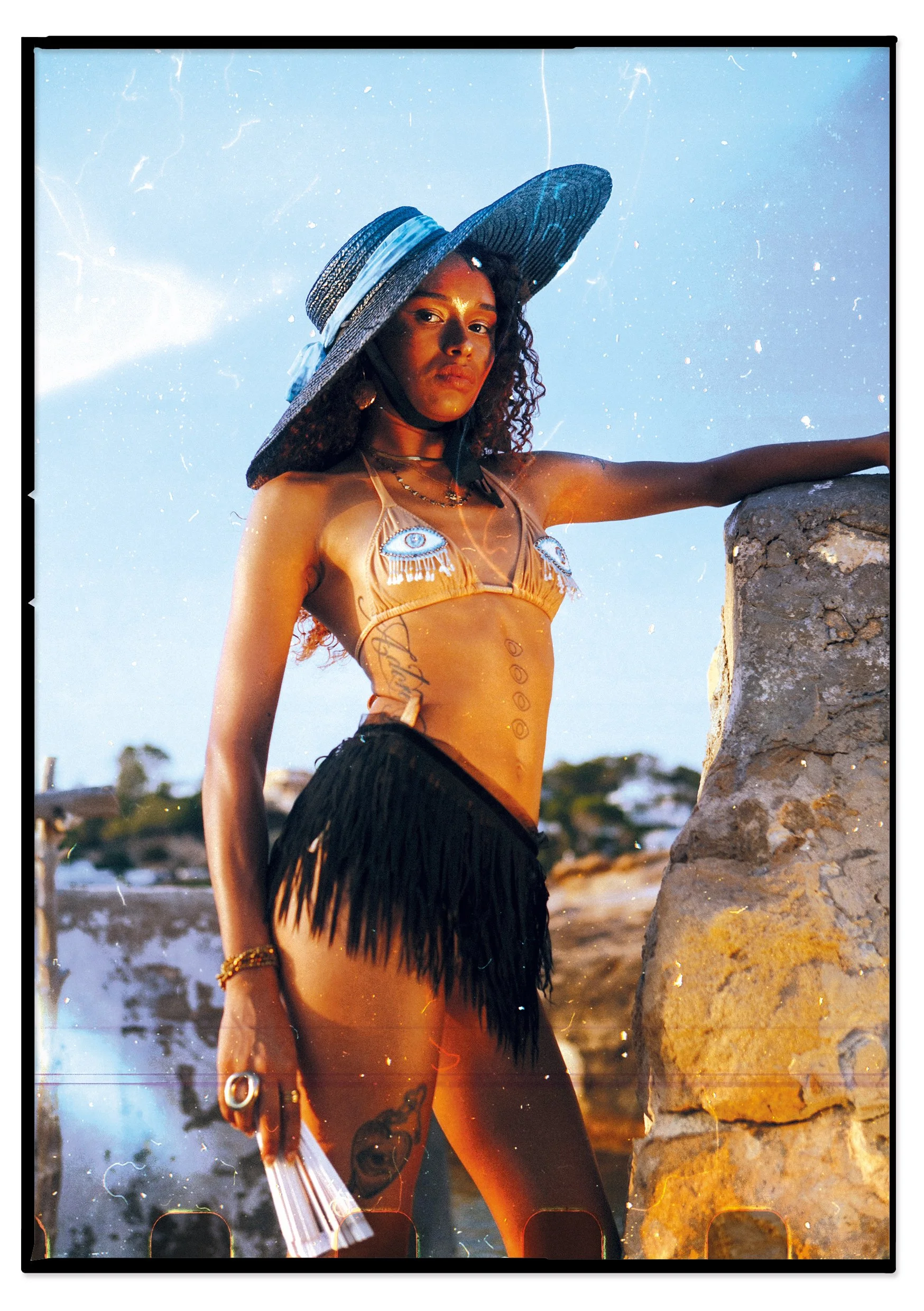 A woman wearing a large sunhat, a bikini top with eye designs, a fringe skirt, and jewelry poses outdoors near a stone structure, with a bright sky and distant landscape in the background.