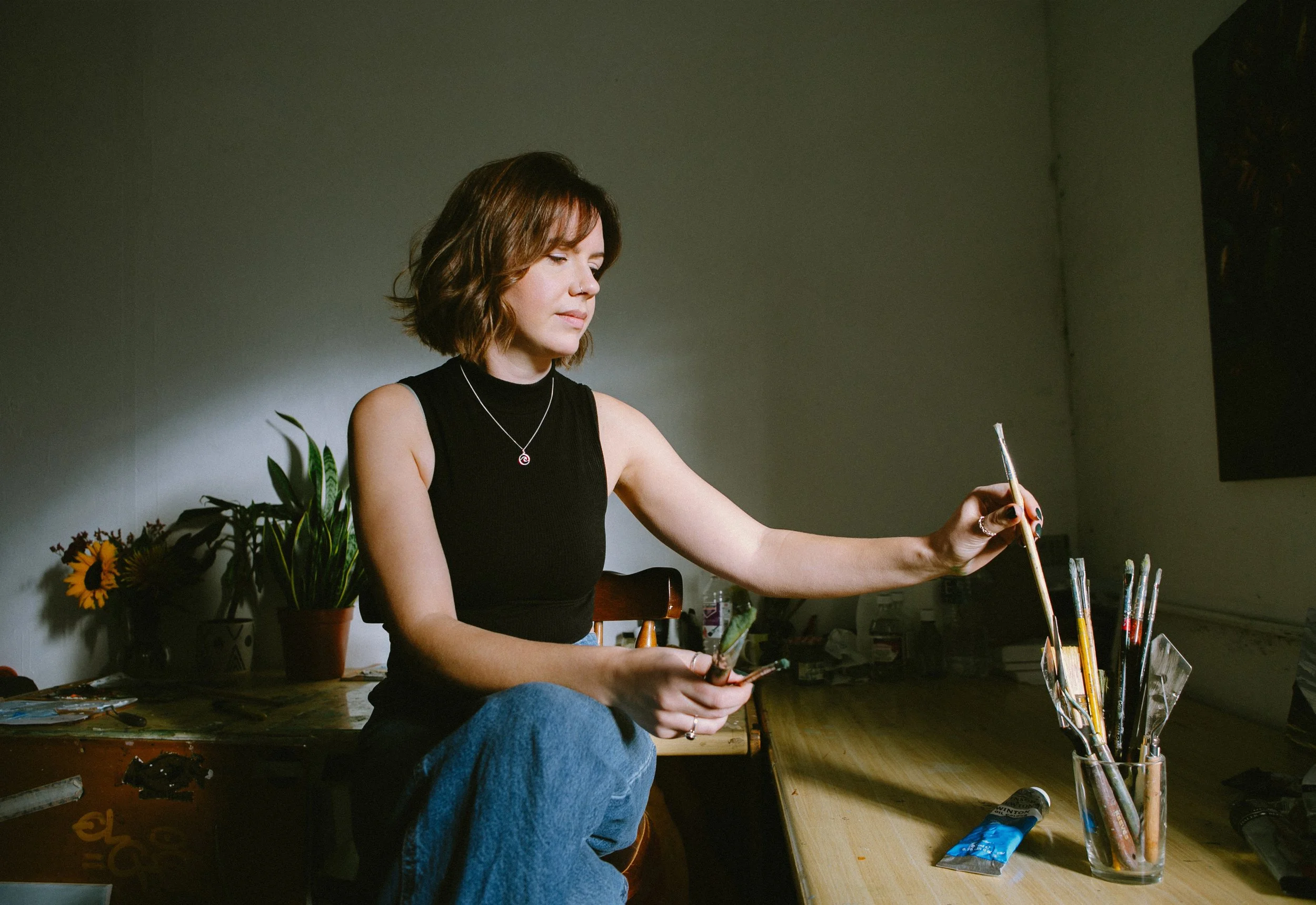 A woman with shoulder-length brown hair wearing a black sleeveless top and blue jeans sitting at a wooden table, holding paintbrushes, with paint supplies and potted plants visible on the table and shelf behind her.