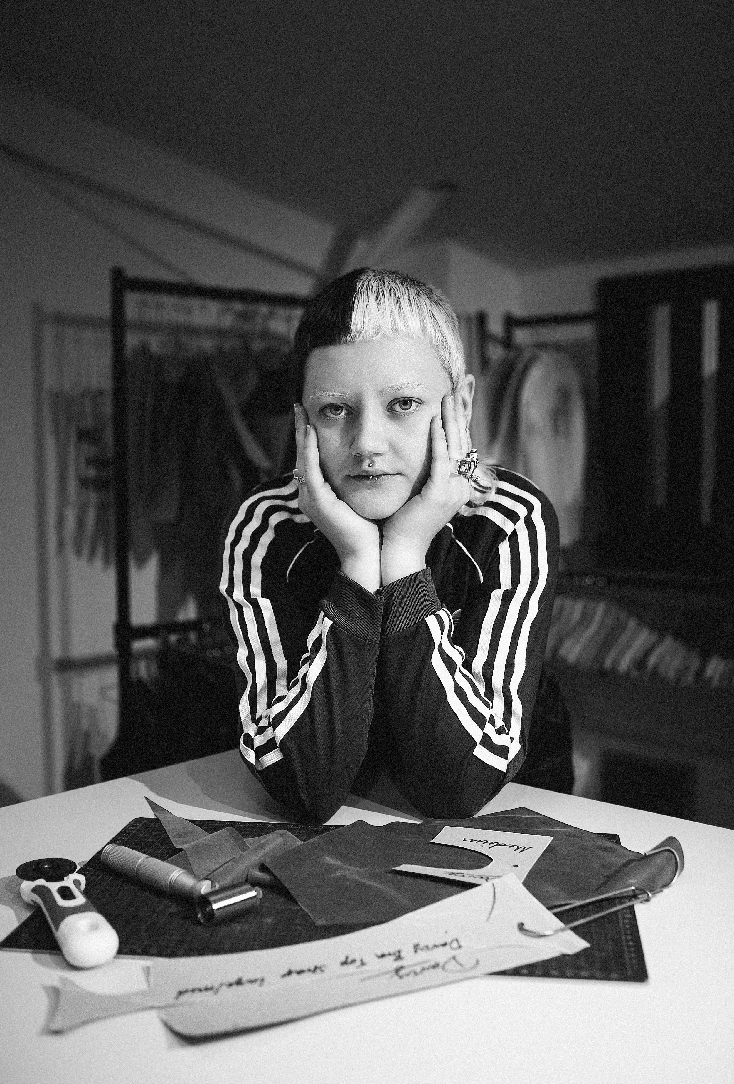 A person with short hair and piercings, sitting at a table with various sewing tools and fabric, resting their face in their hands, in a room with clothing racks in the background.