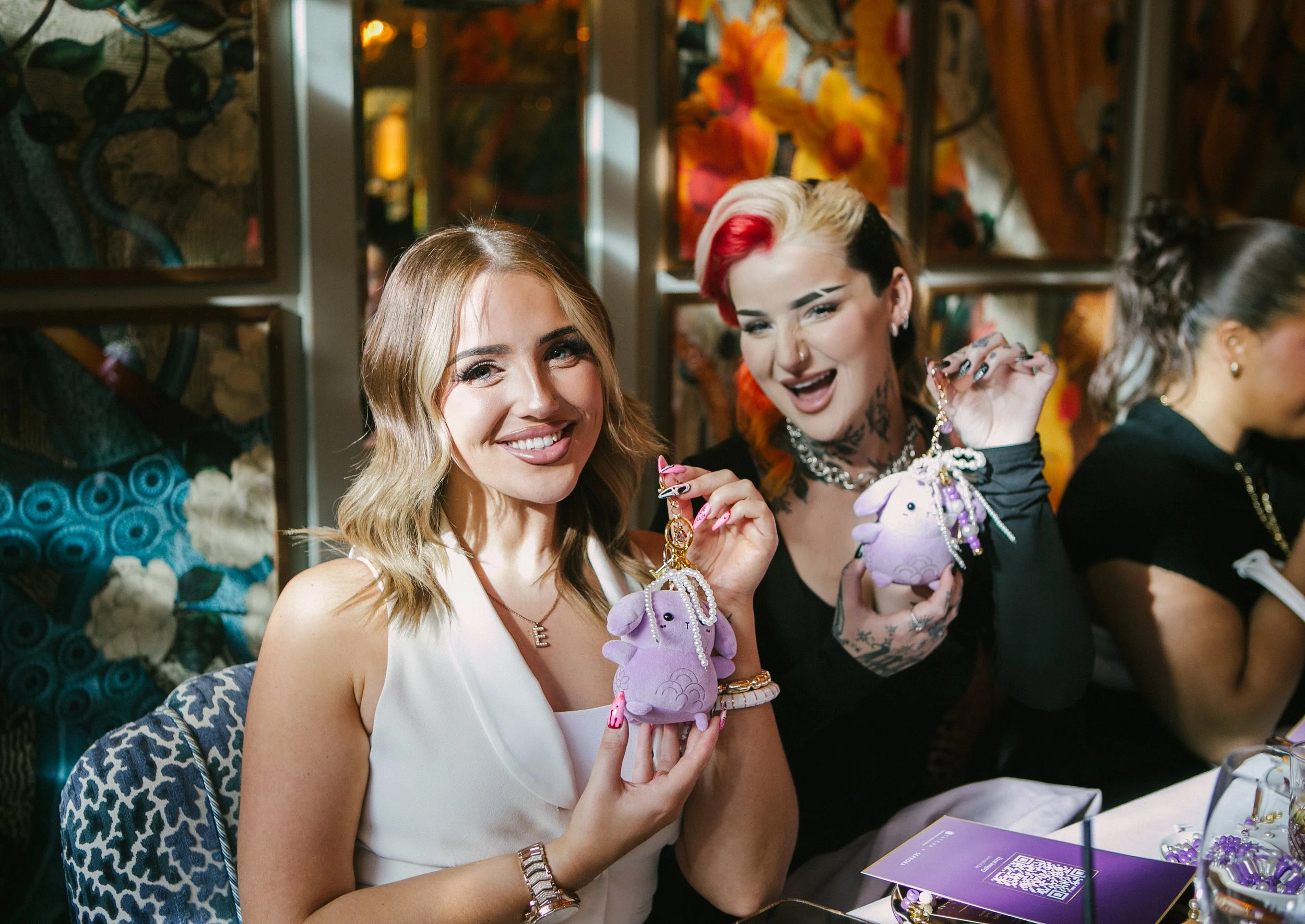 Two women at a table holding purple plush keychains with unicorns, smiling at the camera. The woman on the left has shoulder-length blonde hair, and the woman on the right has short, multicolored hair.
