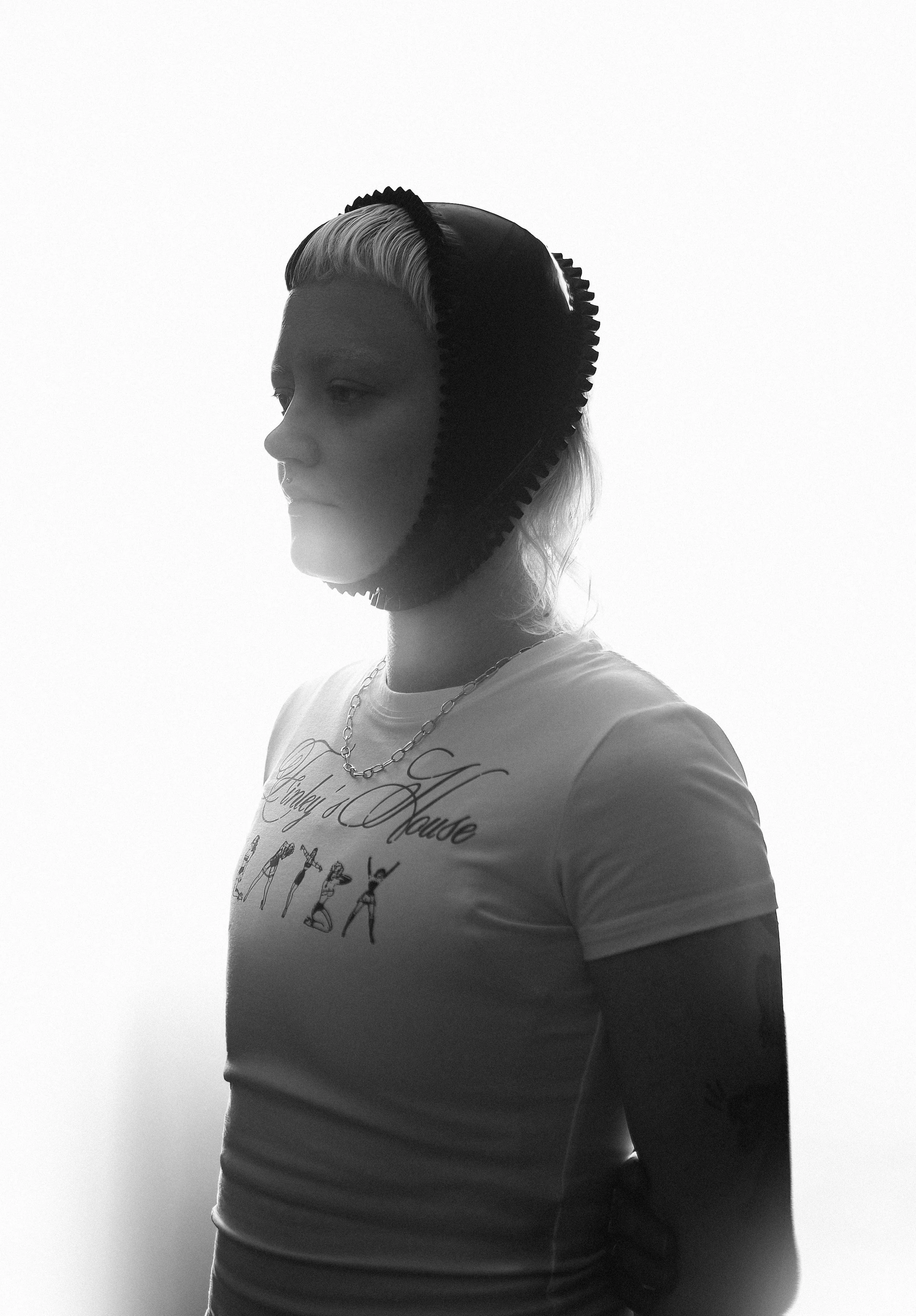 A woman with a blonde ponytail wearing a T-shirt with text and a chain necklace, facing left, with a black foam headband on her head.