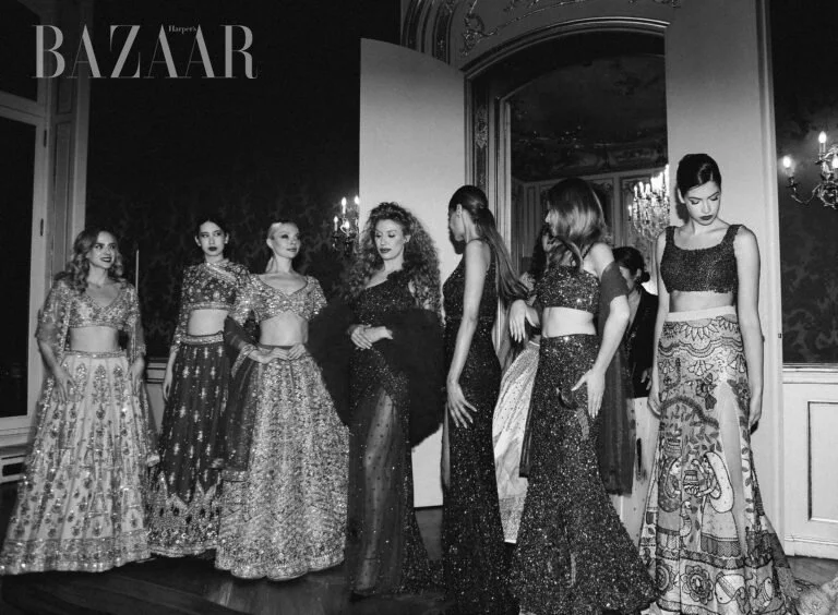 Black and white photo of six women in elaborate, traditional, or formal dresses on a stage or runway, with ornate walls and chandeliers in the background, from Harper's Bazaar magazine.