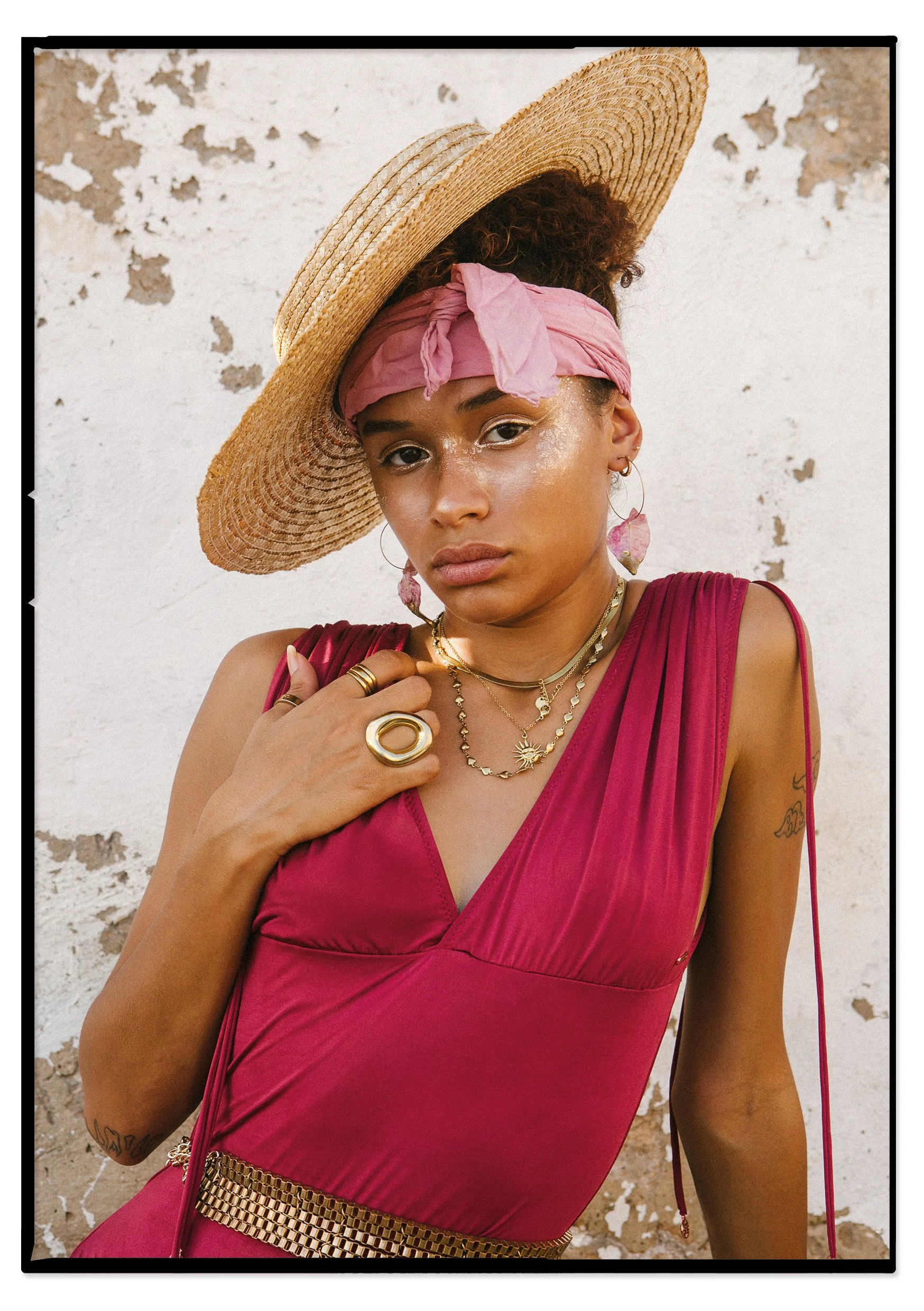A woman wearing a wide straw hat, pink scarf, and a deep pink dress with gold jewelry stands against a weathered, beige and white wall.
