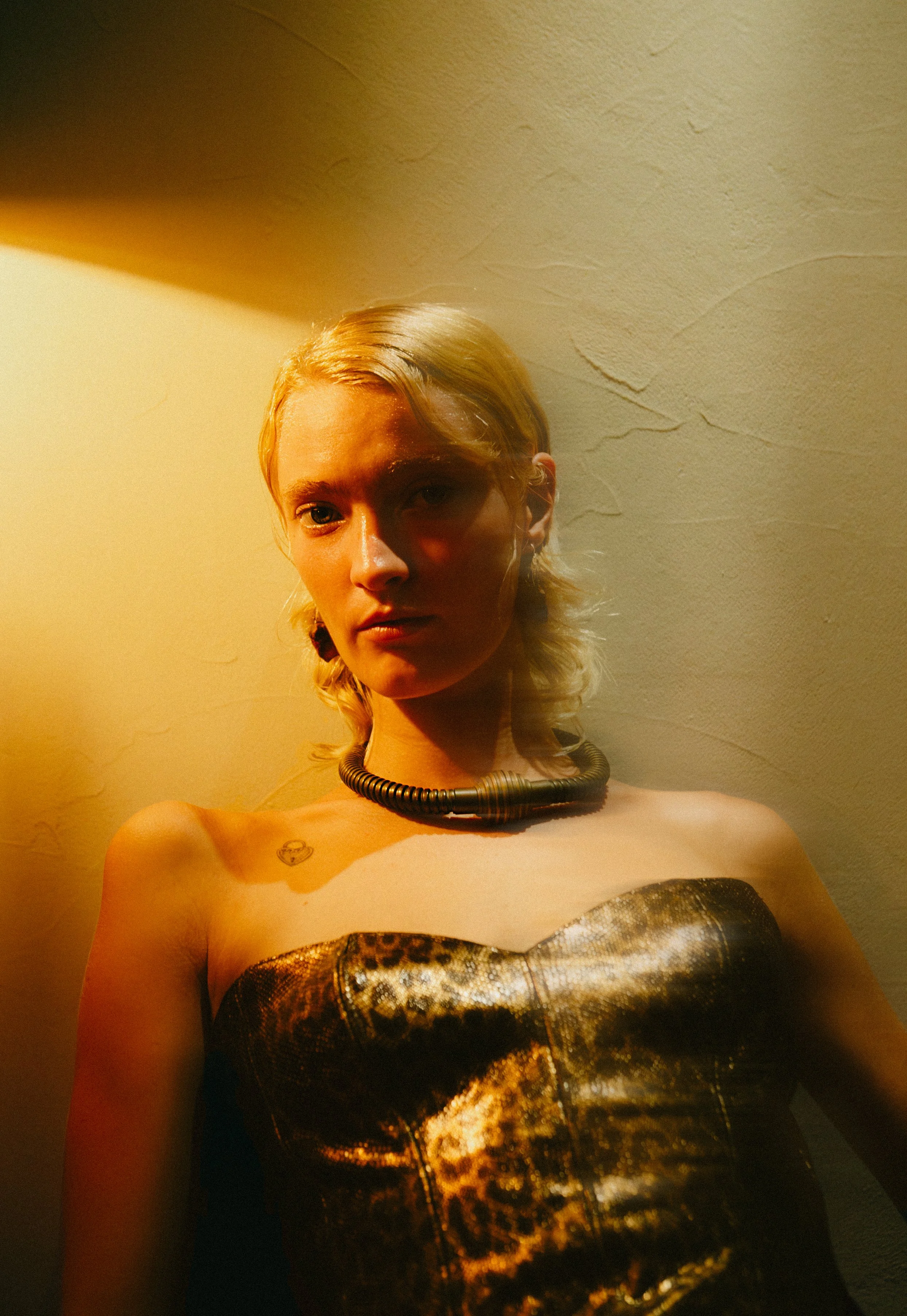 A young woman with blonde hair wearing a strapless dress with a metallic leopard print pattern, accessorized with a chunky necklace and earrings, standing against a textured wall with warm lighting.